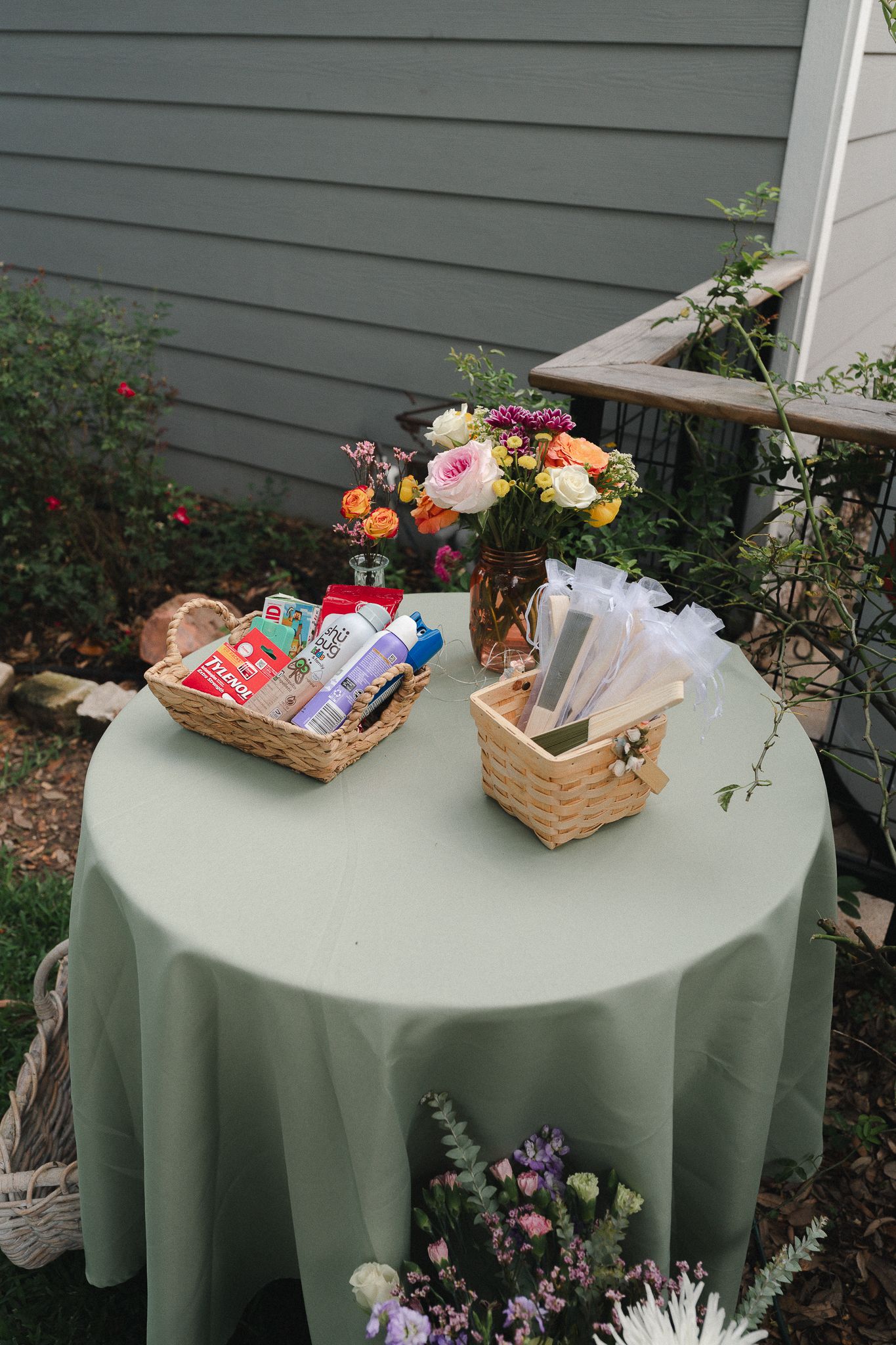 Outdoor round table with sage green tablecloth, wicker baskets, and colorful floral arrangement against gray siding.