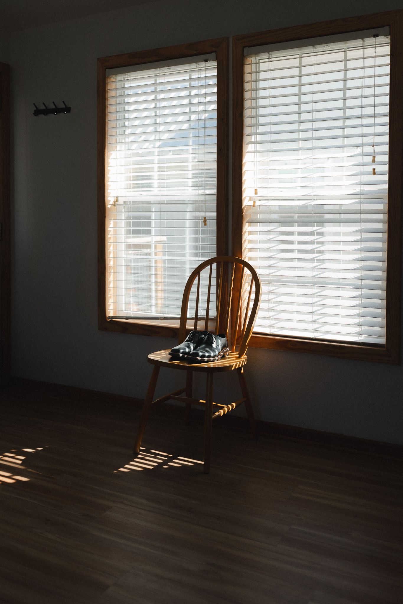 Wooden Windsor chair sits in sunlit room with venetian blinds casting shadows on hardwood floor.