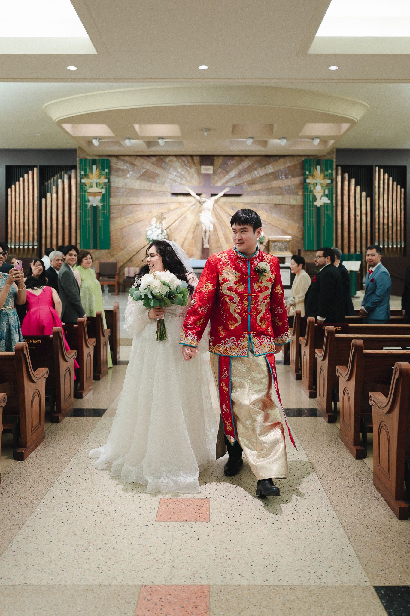 Bride and groom walk down church aisle, groom in red Chinese dragon jacket, bride in white gown.