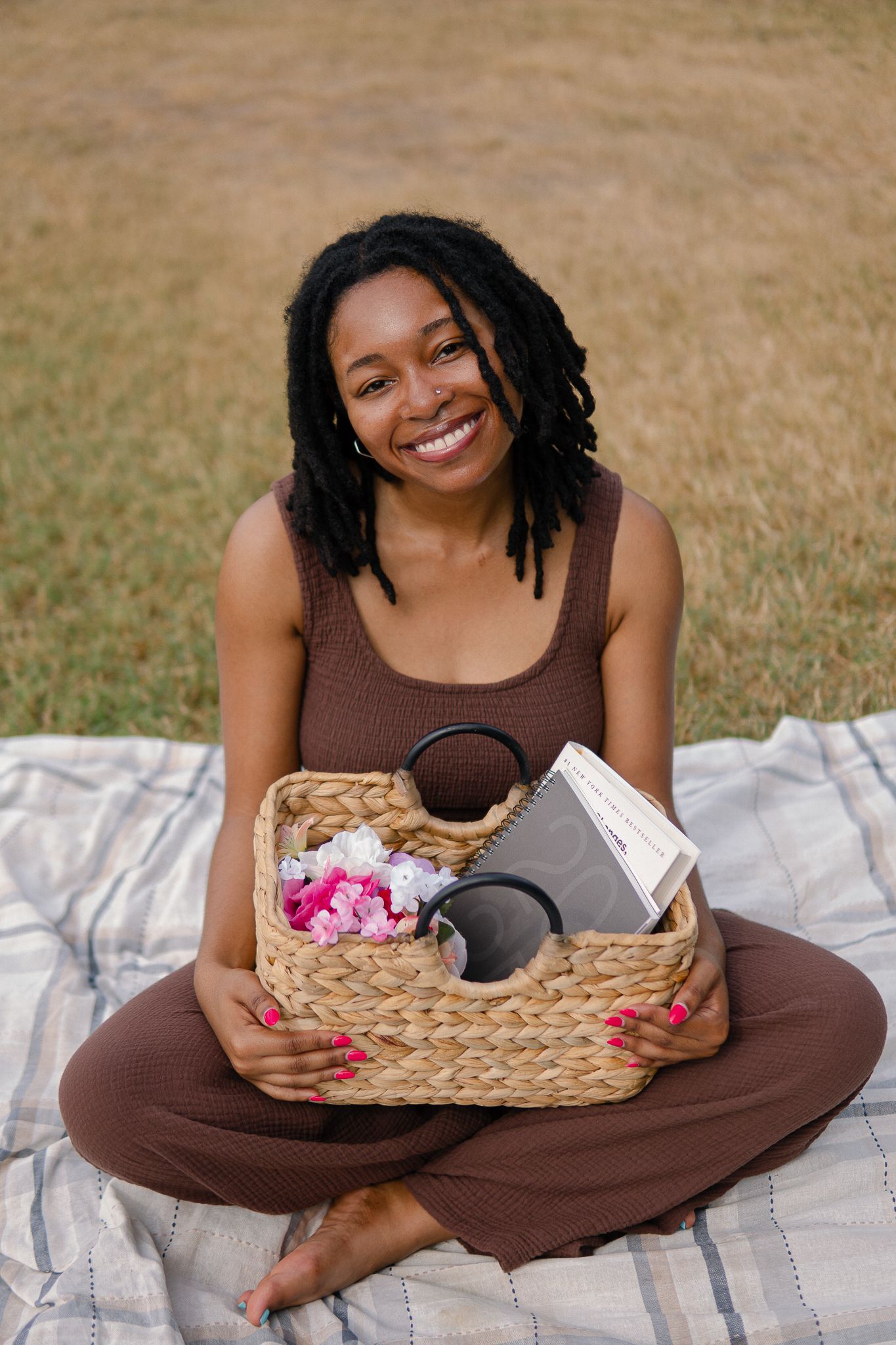 Person in brown dress sits cross-legged on picnic blanket with woven basket of flowers in a sunny outdoor setting.