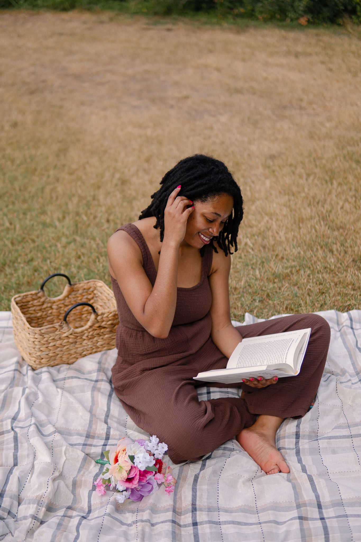 Someone in a brown dress relaxes on a picnic blanket while reading a book in a grassy field.