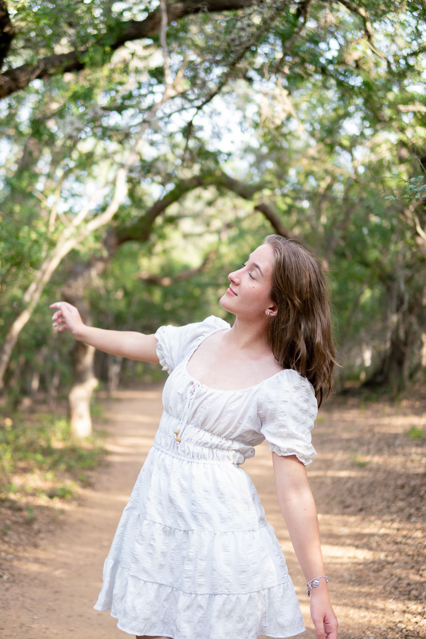 A person in a flowing white dress twirls joyfully on a tree-lined dirt path during golden hour.