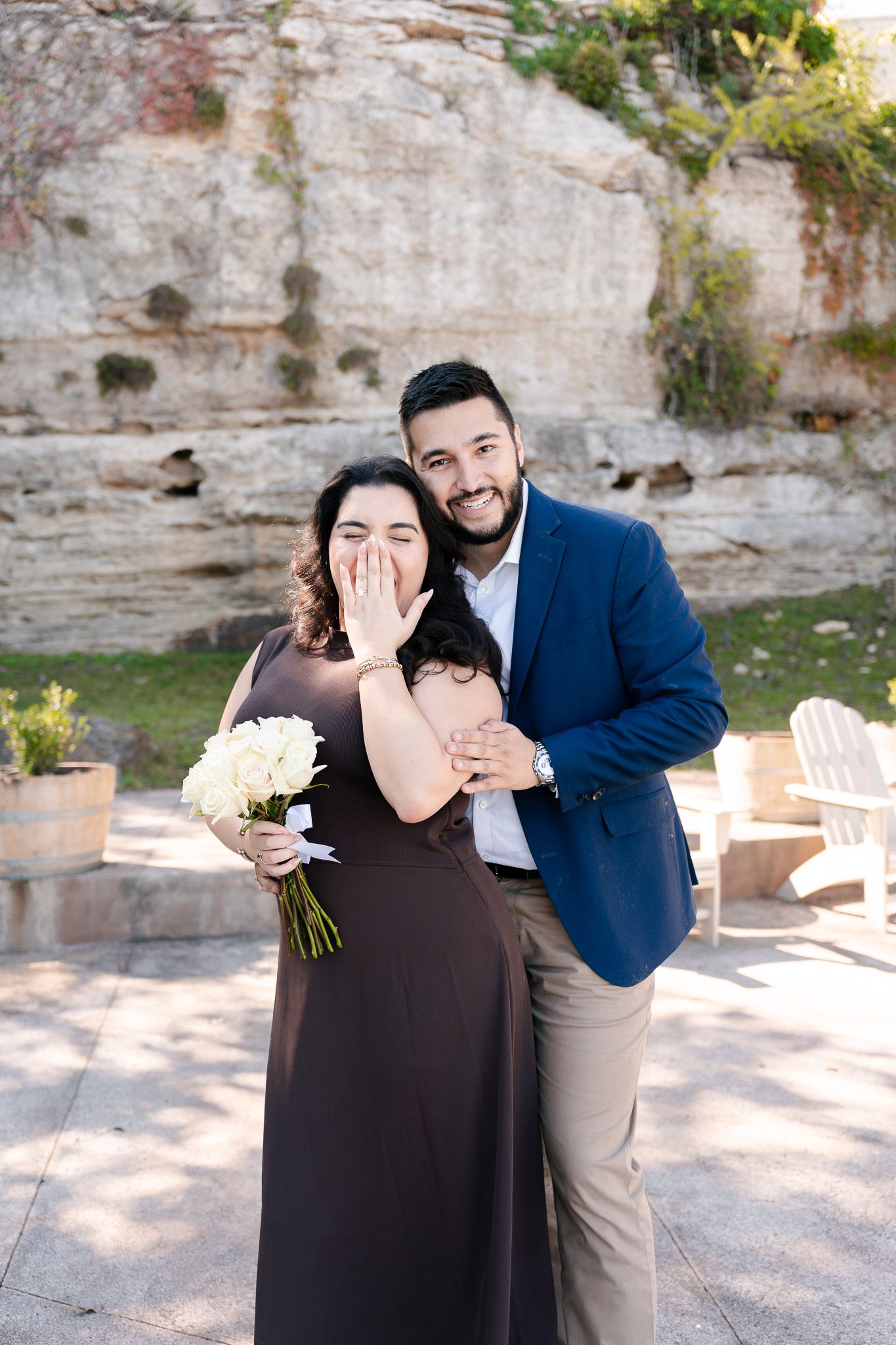 A couple shares a joyful moment at an outdoor venue with stone walls and potted plants, the woman holding white flowers.