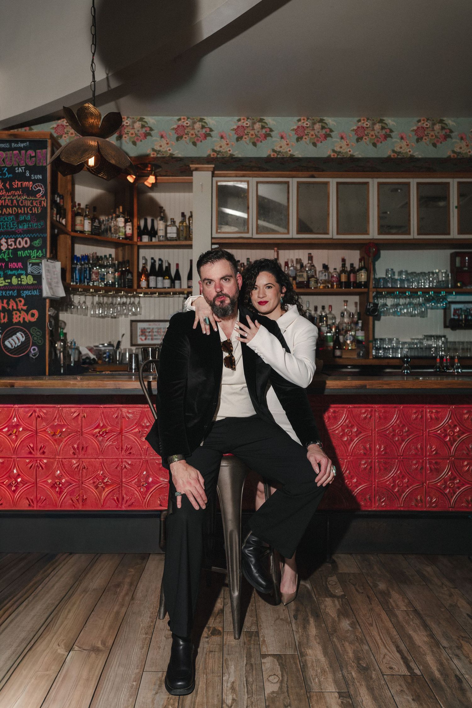 A stylish couple poses together at a vintage bar, man seated in black suit, woman in white blazer leaning close.