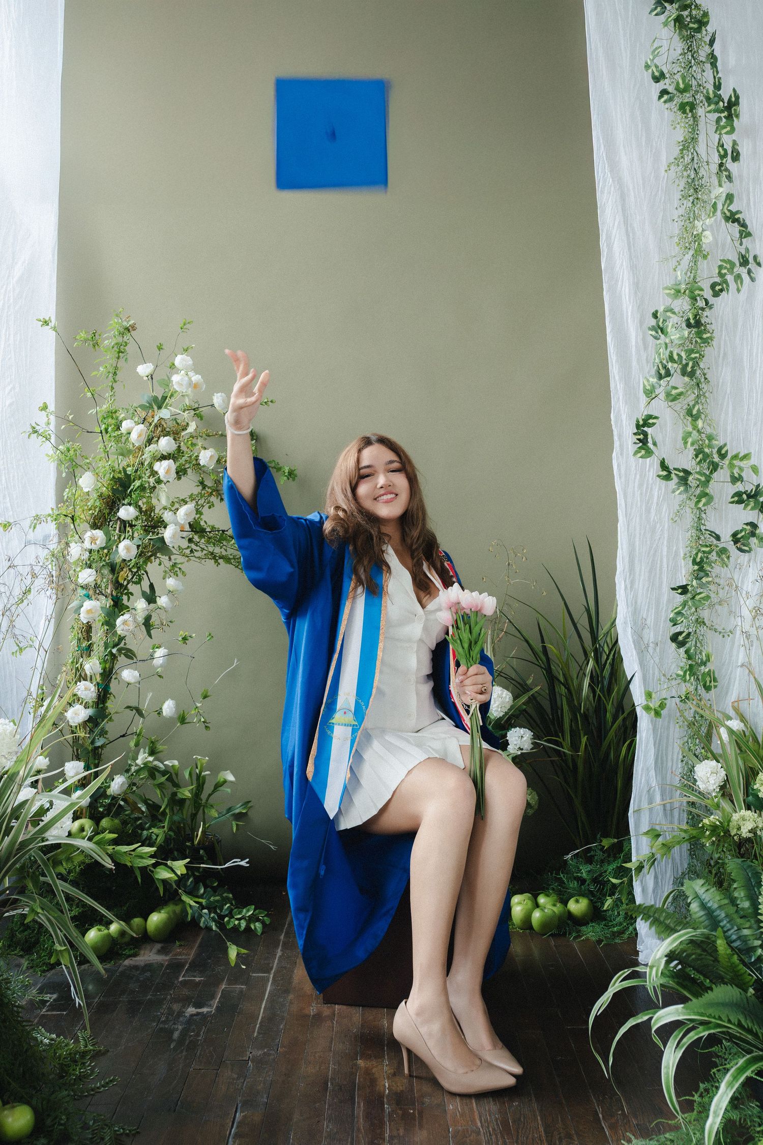 Smiling graduate in blue cap and gown sitting with flowers, surrounded by greenery in a decorated photo setting.