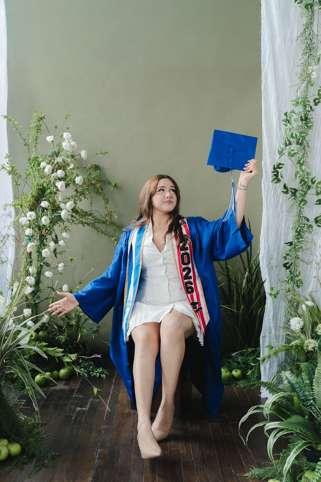 Woman in blue graduation gown and cap sitting among white flowers and green plants in a studio setting.