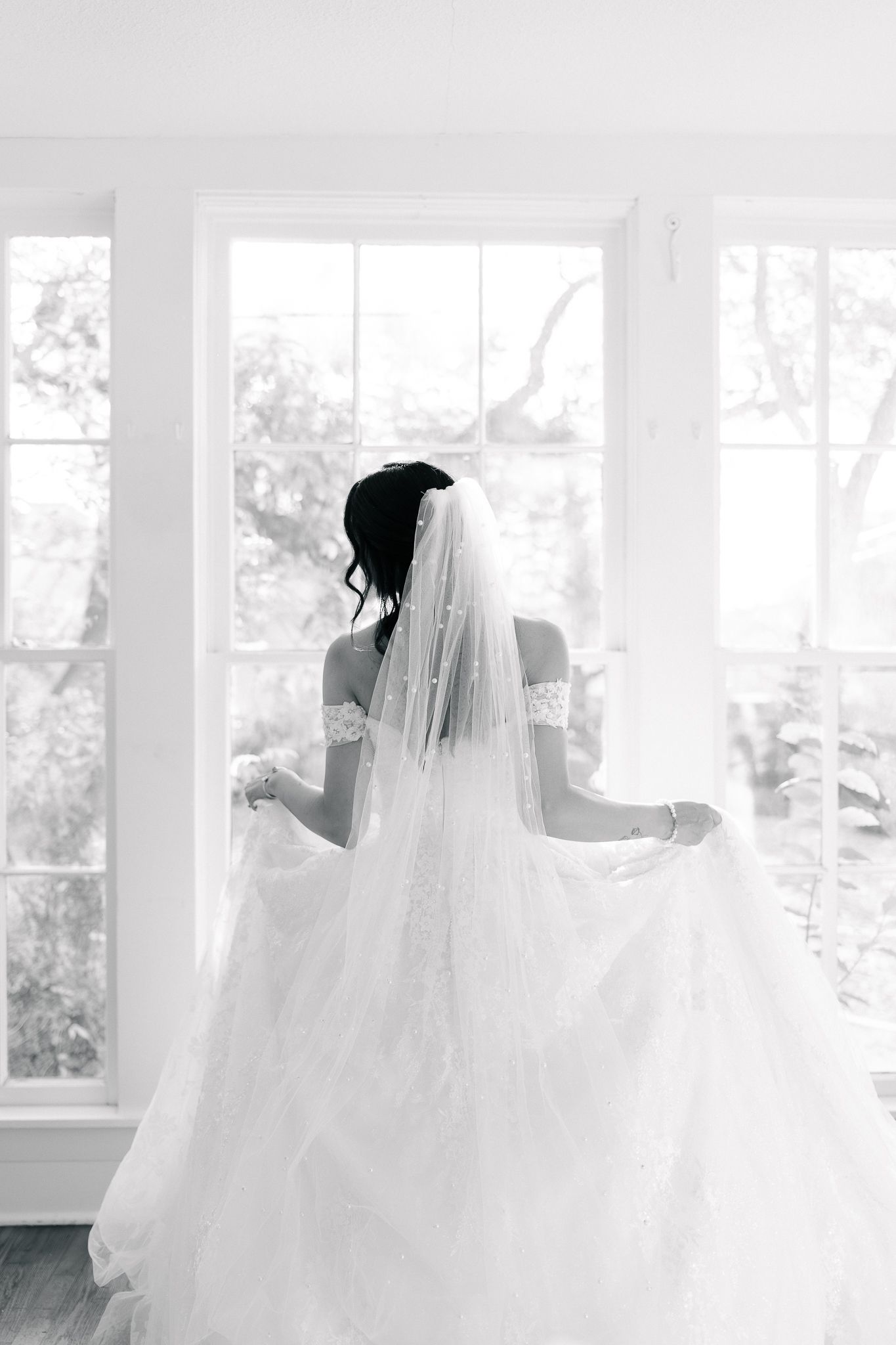 A bride in a flowing white gown twirls in an elegant room filled with natural light from large windows.