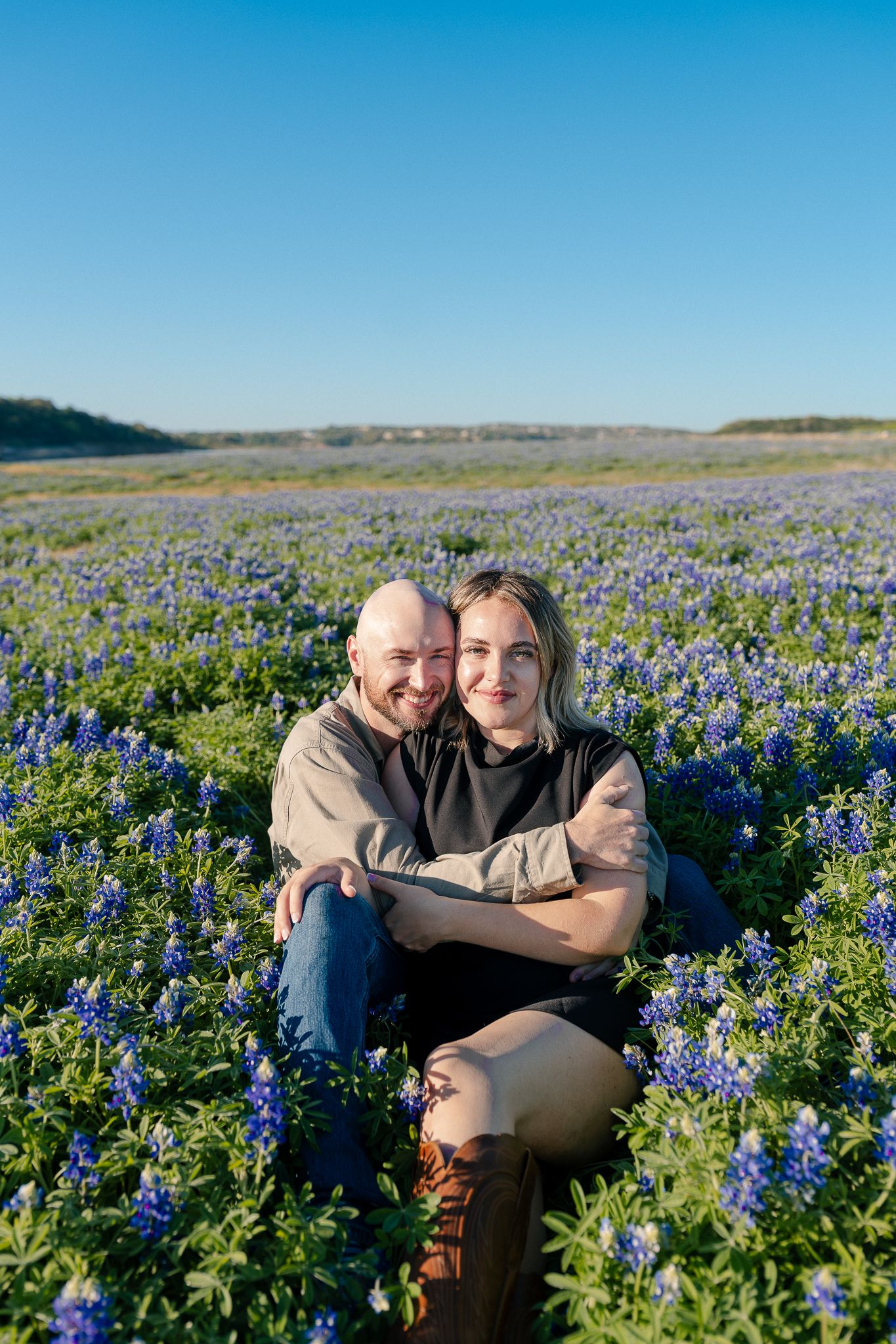 A couple sits together in a vibrant field of blooming Texas bluebonnets under a bright blue spring sky.