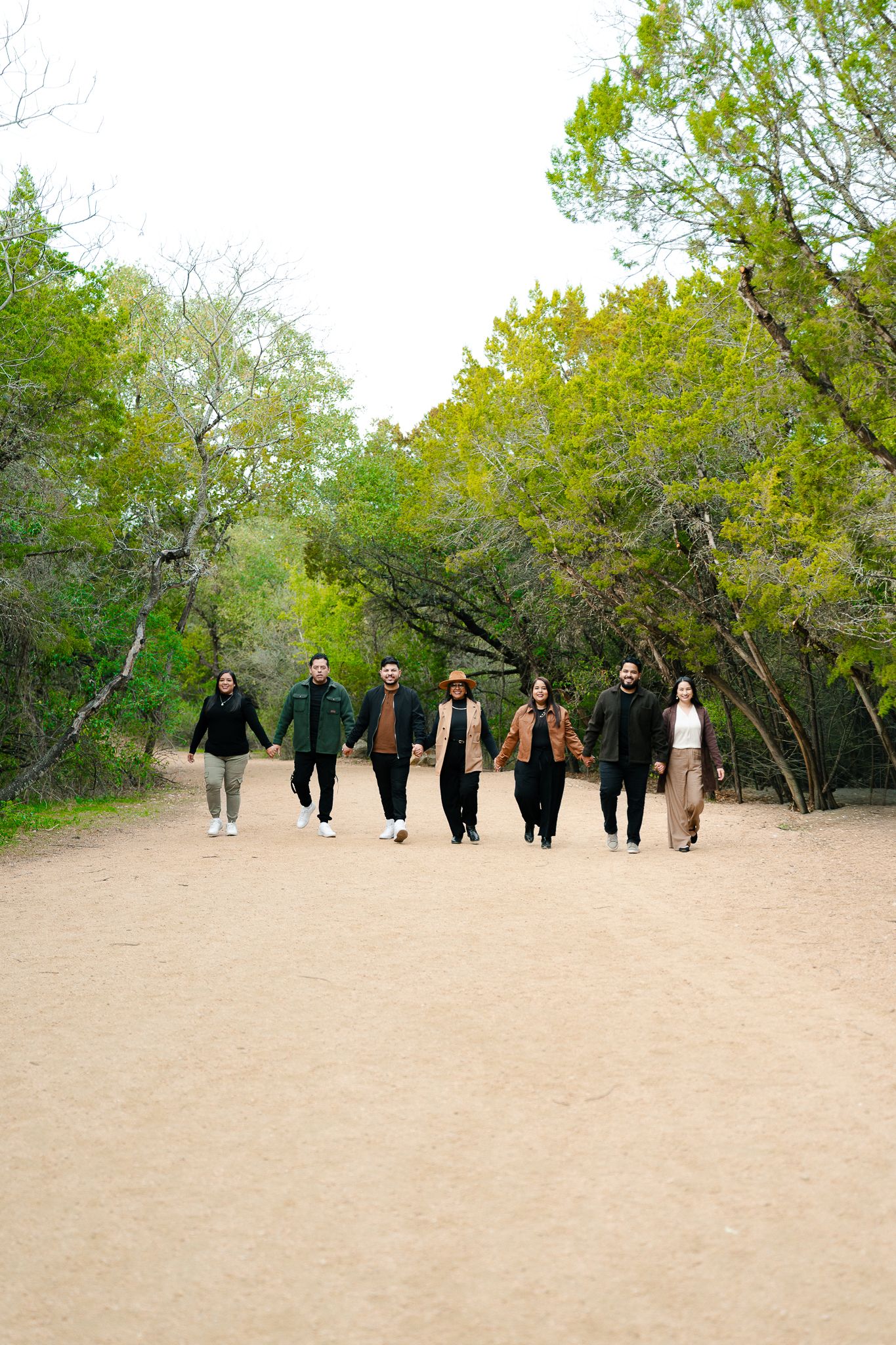 A group of people in dark clothing walk together through a sandy path lined with green trees.