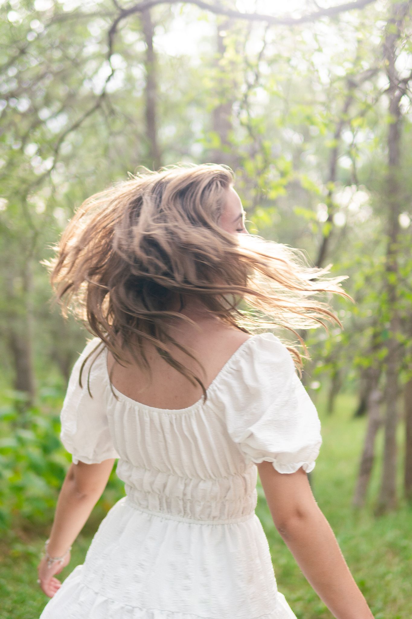 A person in a flowing white dress spins through a sunlit forest creating a dreamy, ethereal scene.