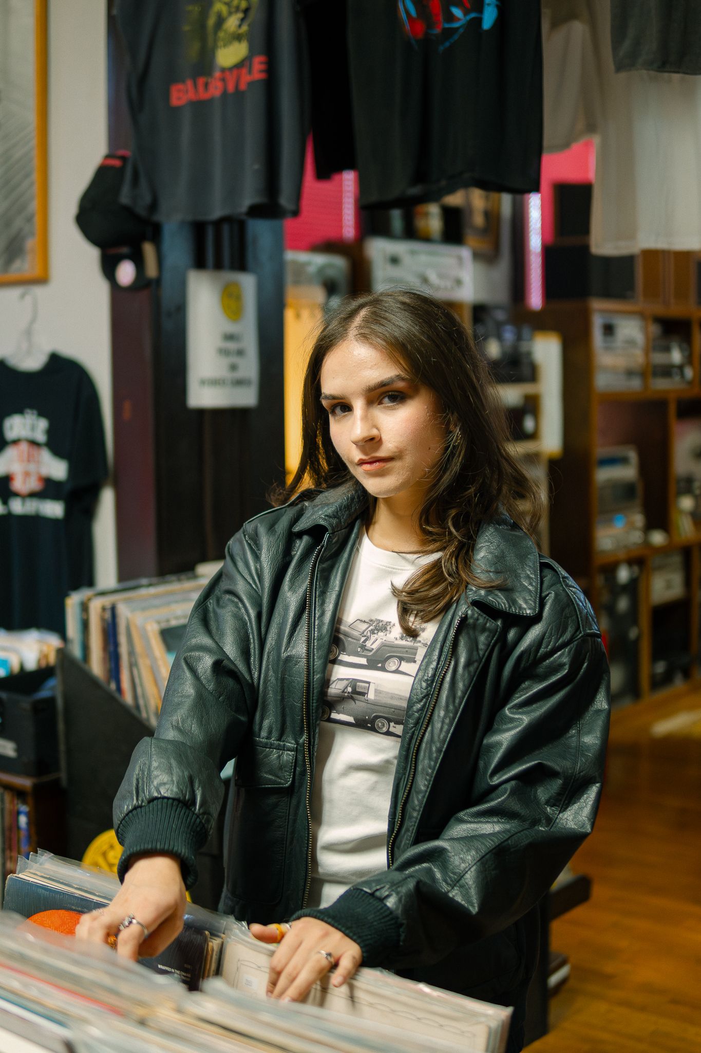 Person in leather jacket browses through vinyl records at a music store with t-shirts displayed on wall.