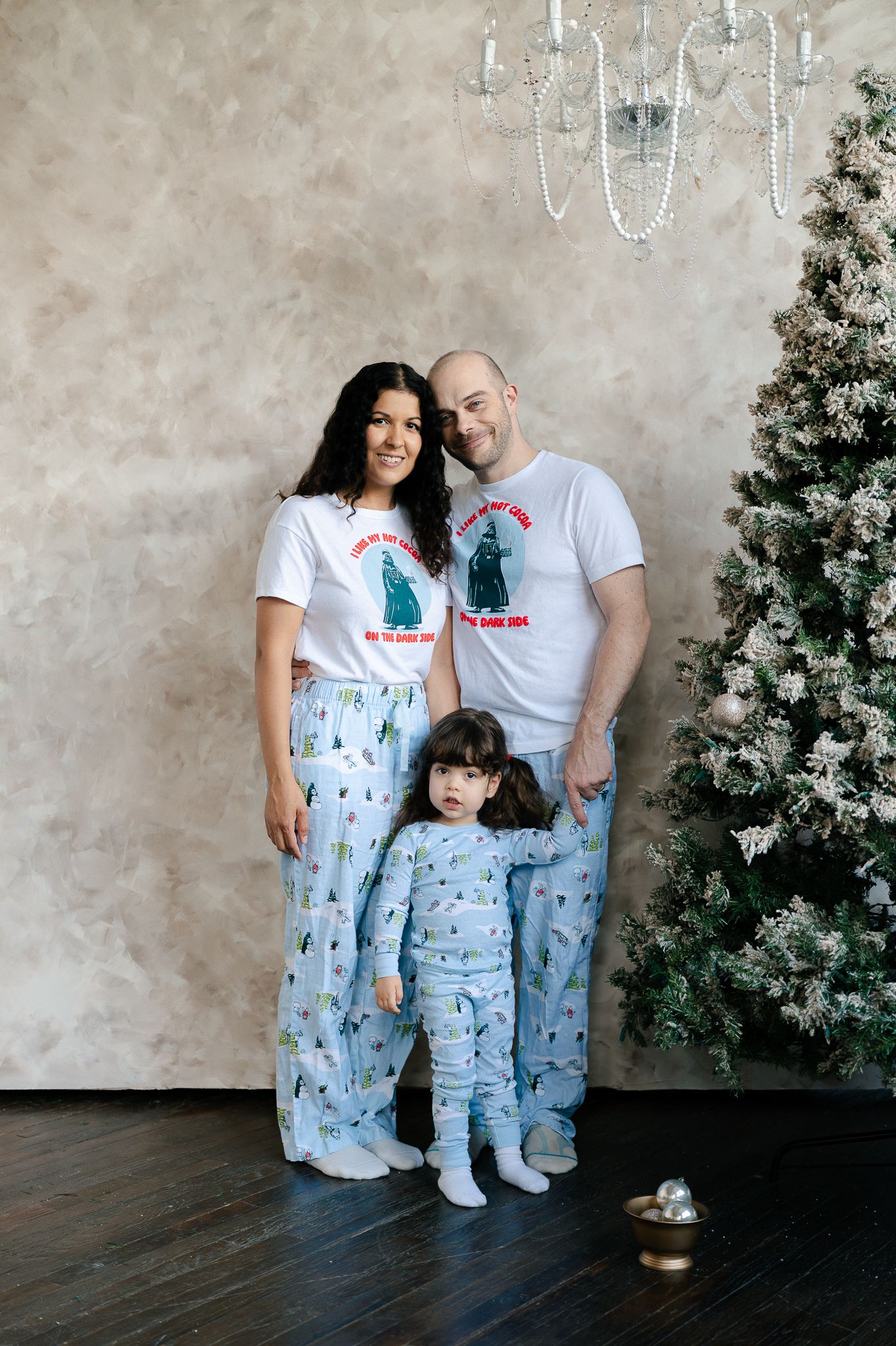 A family wearing matching dinosaur-themed pajamas poses together near a Christmas tree and chandelier.