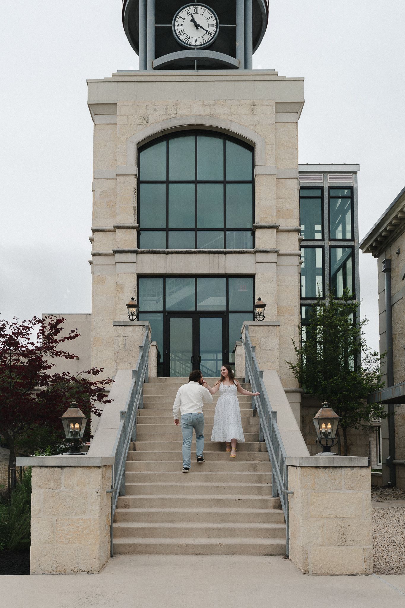 Couple in wedding attire walking up stone steps toward a clock tower building with large windows.
