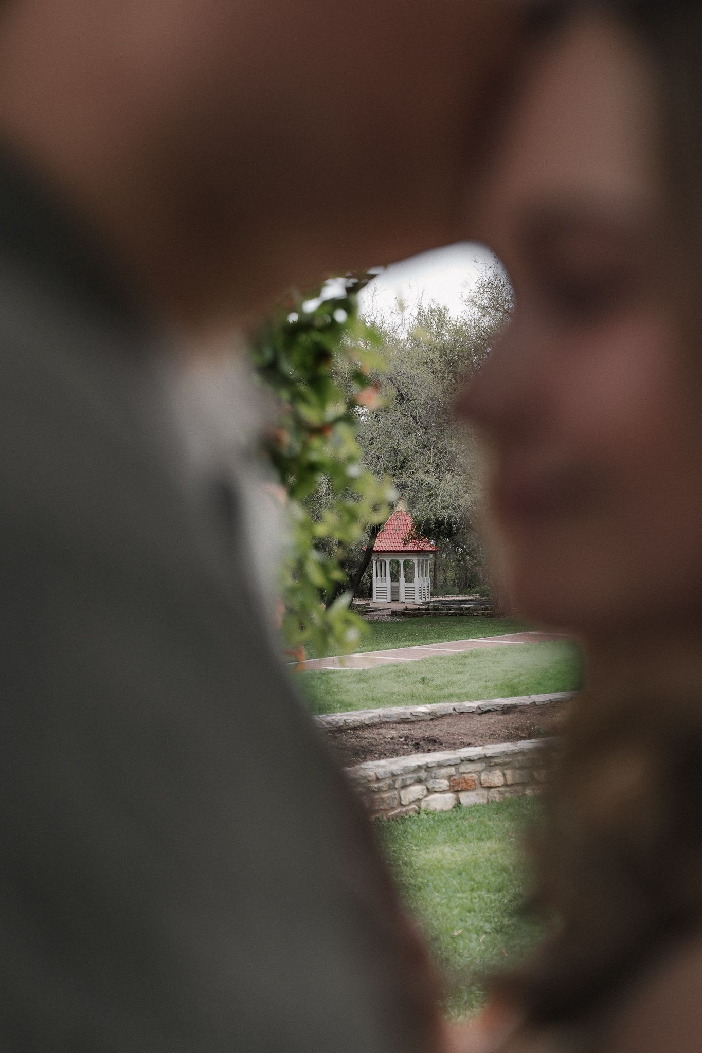 Blurred couple framing a charming garden gazebo with red roof surrounded by lush greenery in soft focus.