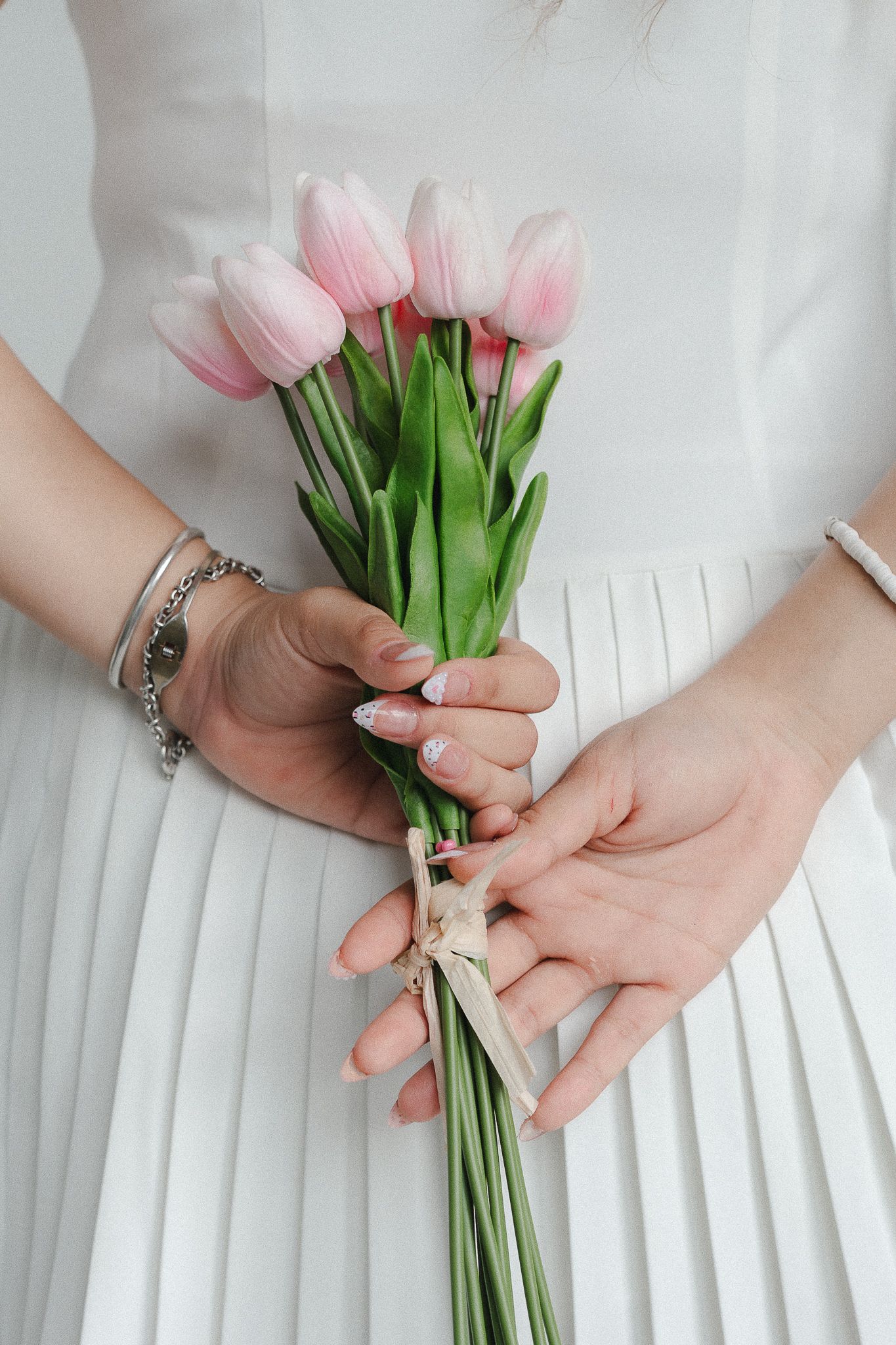 Bride in white pleated dress holding a bouquet of pink tulips tied with ribbon, showcasing elegant manicure and silver bracelet.