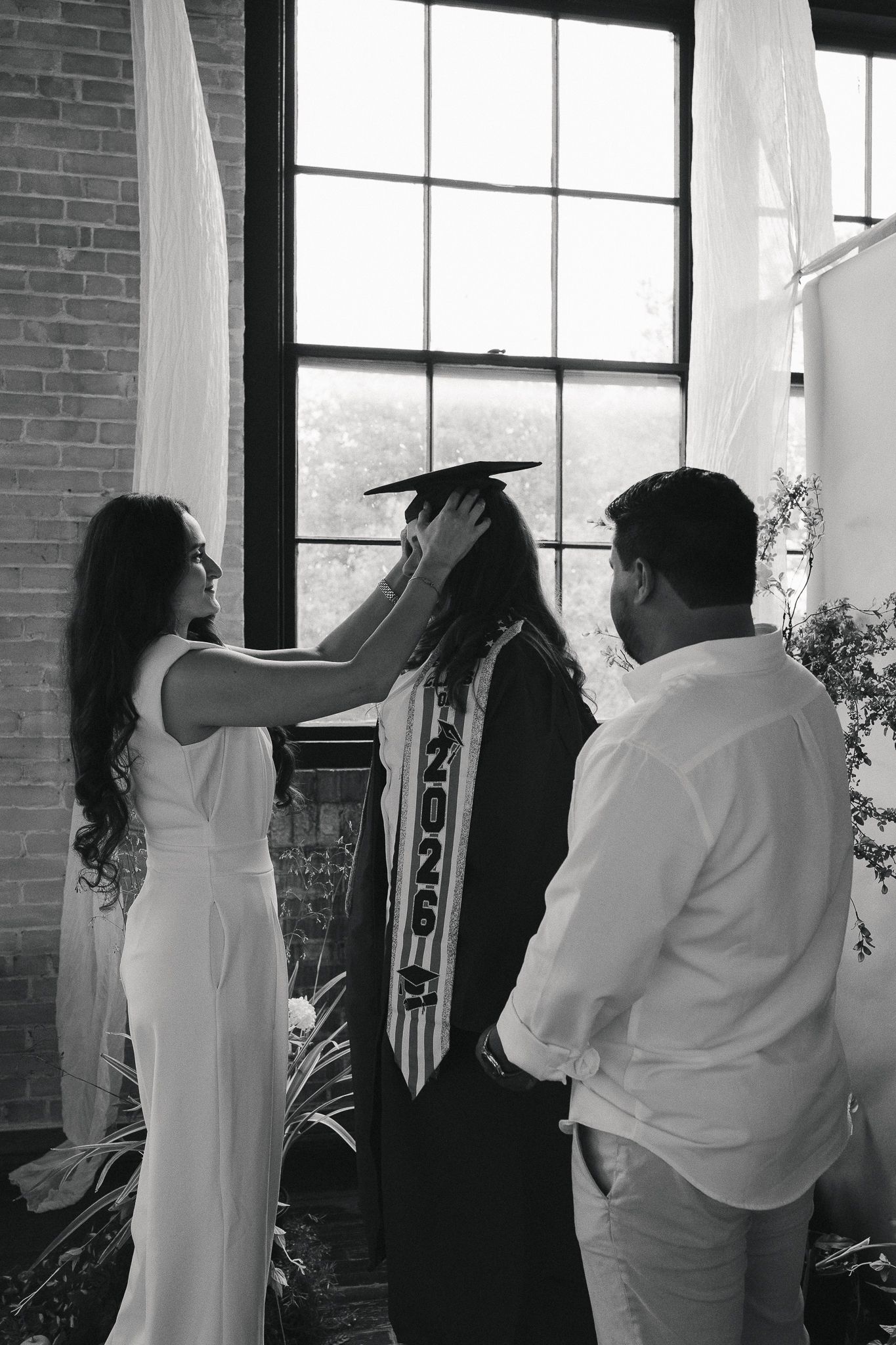 Black and white photo of a graduate in cap and gown with family adjusting their graduation attire indoors.