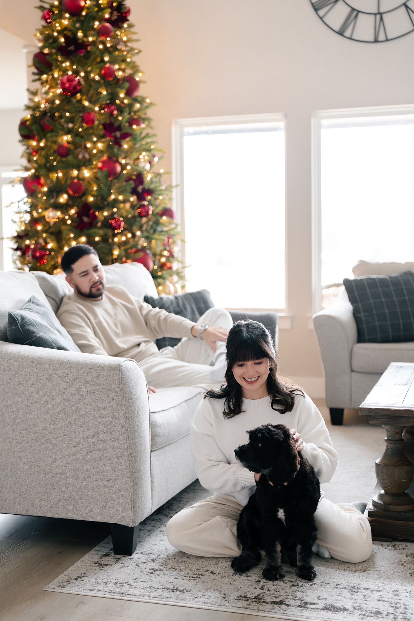 A cozy living room scene with a decorated Christmas tree, gray couch, and black dog on a patterned rug.