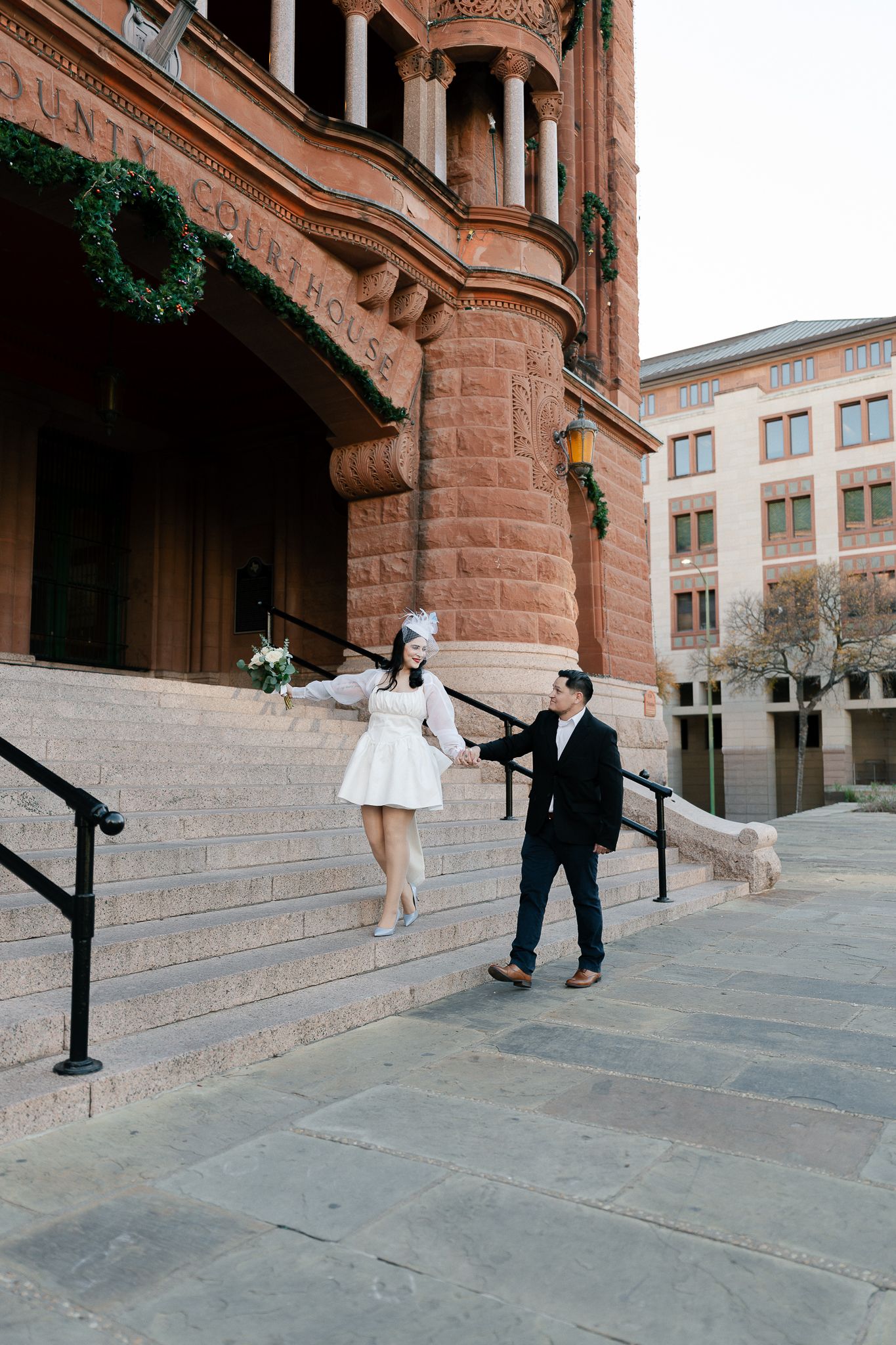 A couple dances playfully on the steps of a historic red sandstone building with ornate architectural details.