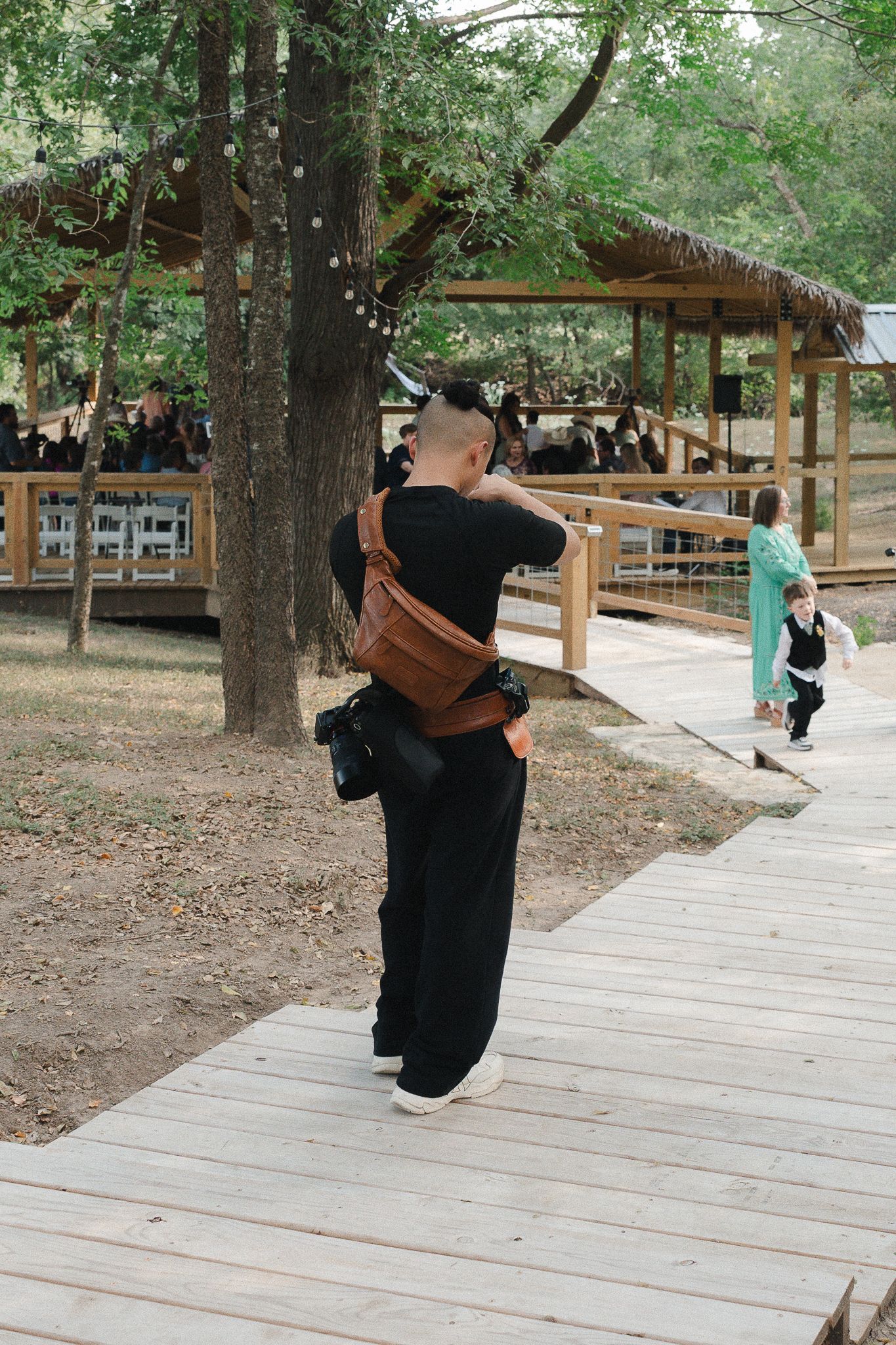 Photographer in black outfit with brown bag captures shots on wooden boardwalk path surrounded by trees and pavilion.