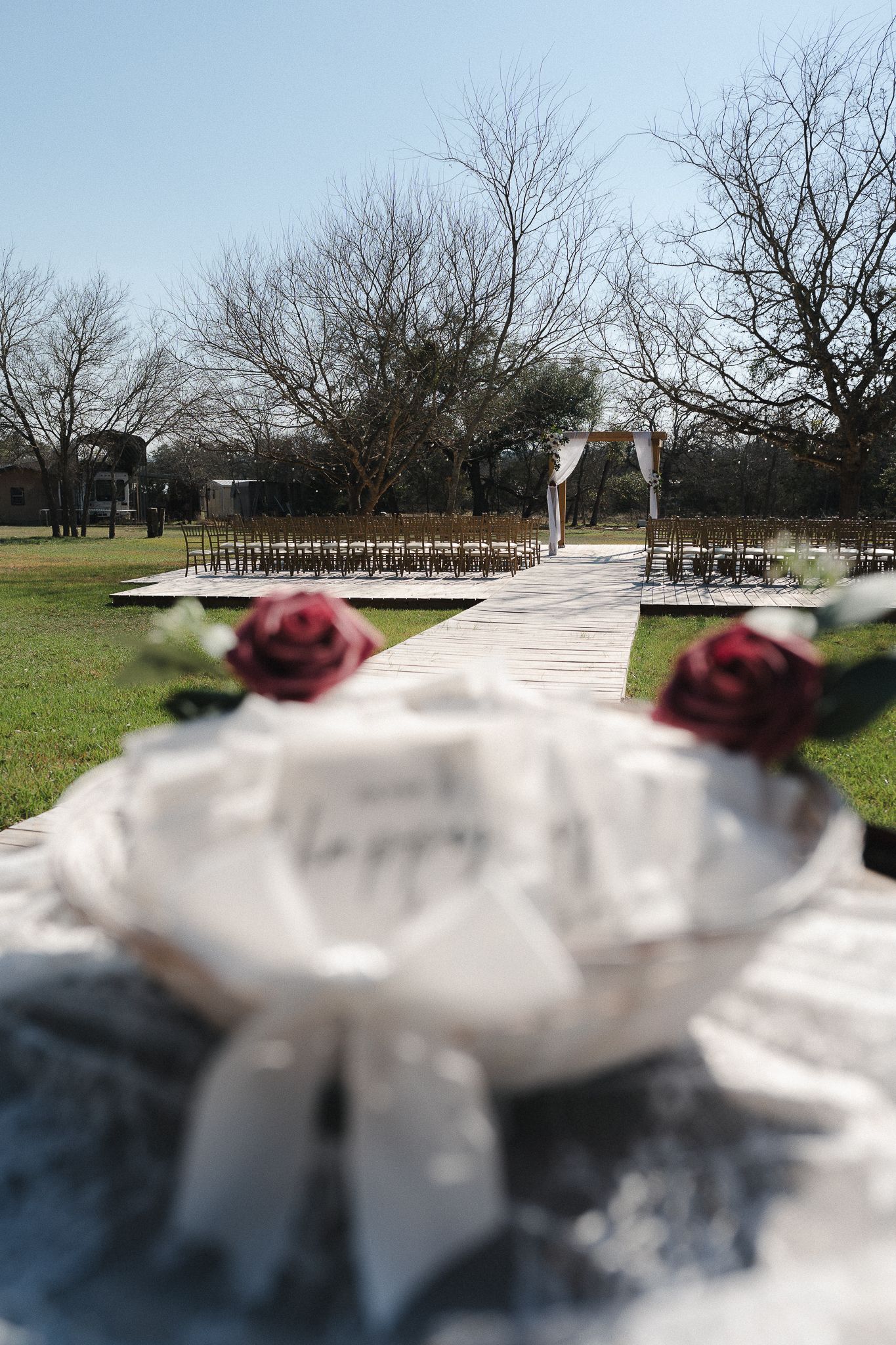 Outdoor wedding ceremony aisle with rose decorations, wooden walkway, chairs, and floral arch amid bare trees on a sunny day.