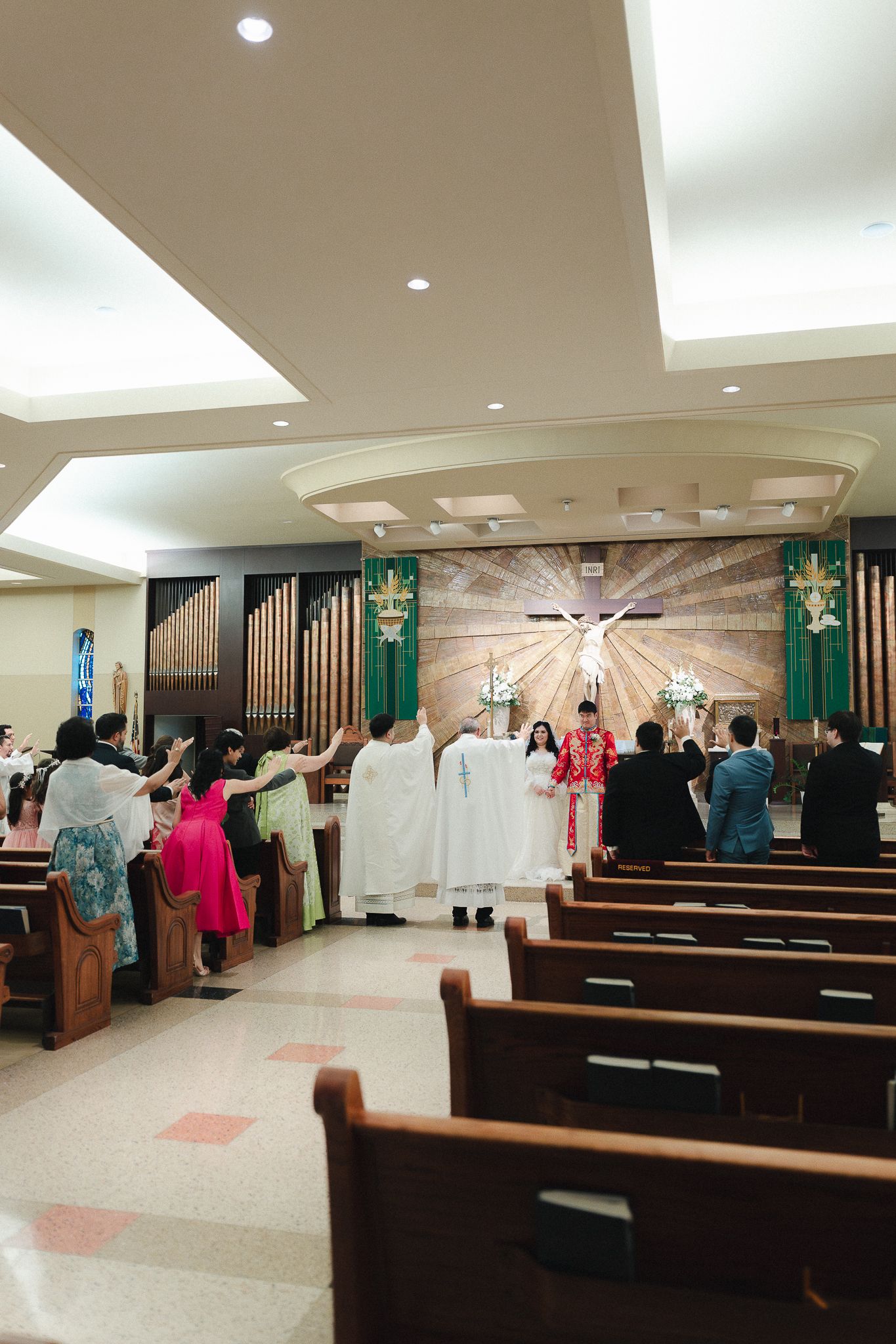 Catholic wedding ceremony at church altar with priest, couple, and guests raising hands in prayer.