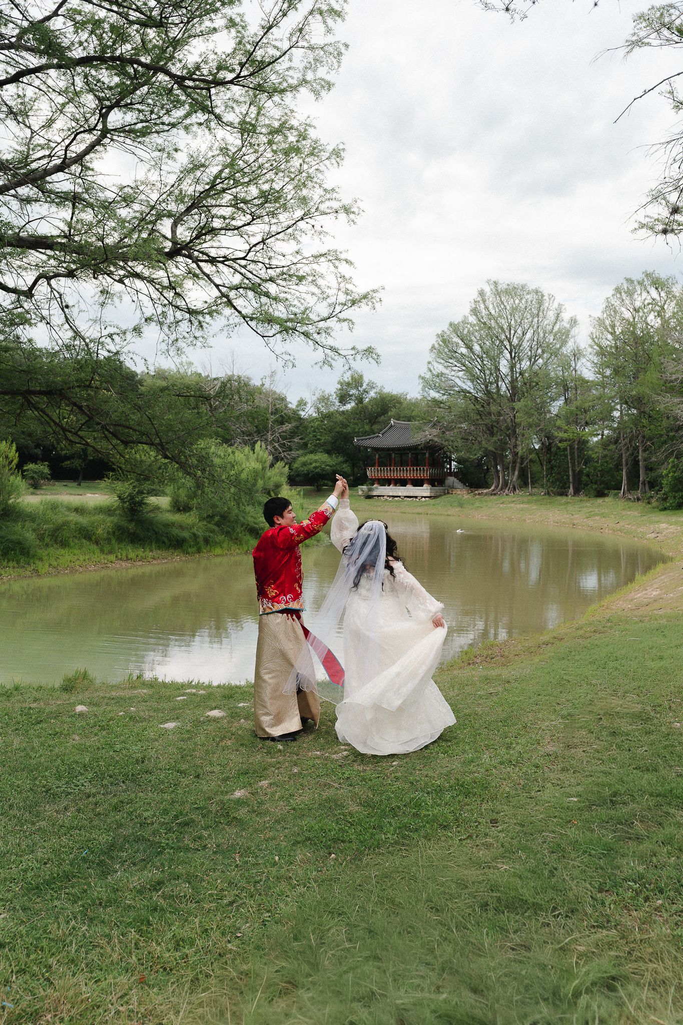 Couple in traditional Korean hanbok attire dancing by a serene pond with a pavilion in a lush green park.