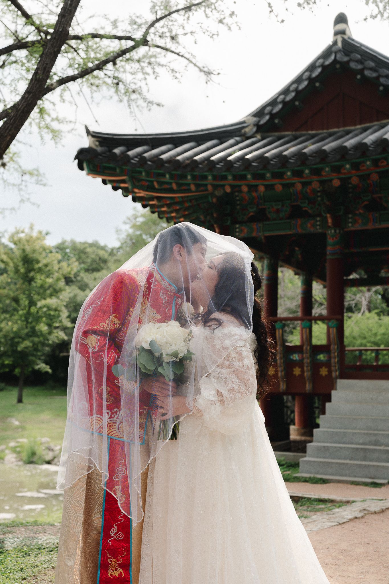 Couple kissing under bridal veil in traditional Korean wedding attire near a pagoda.