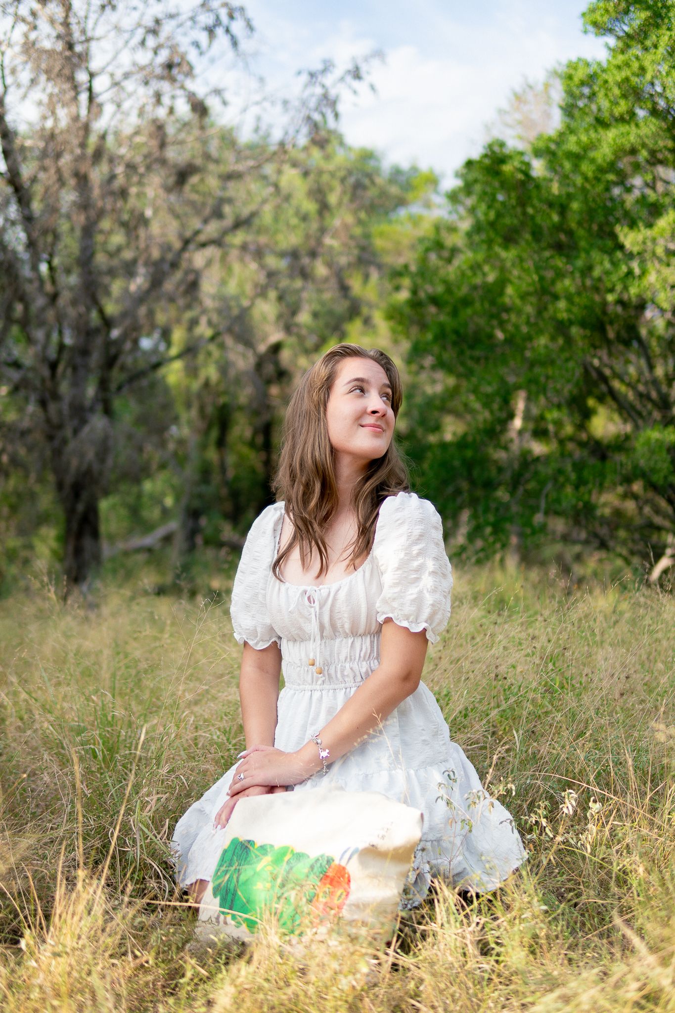 A figure in a white dress sits thoughtfully in a sunlit meadow of tall grass surrounded by trees.