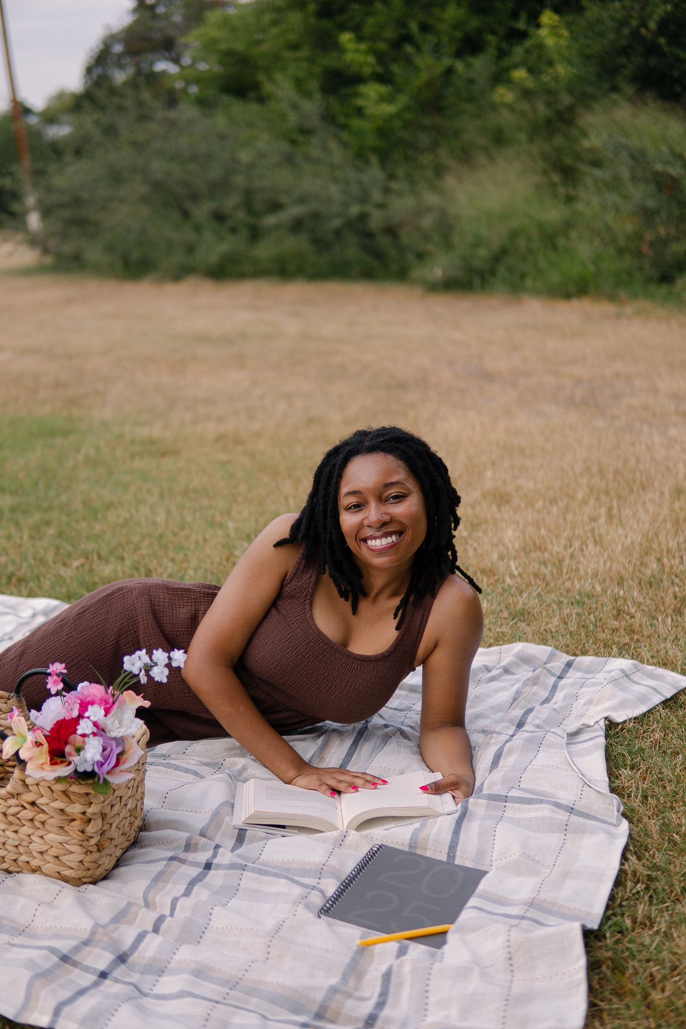A person lies on a picnic blanket in a grassy field with a basket of flowers on a sunny summer day.