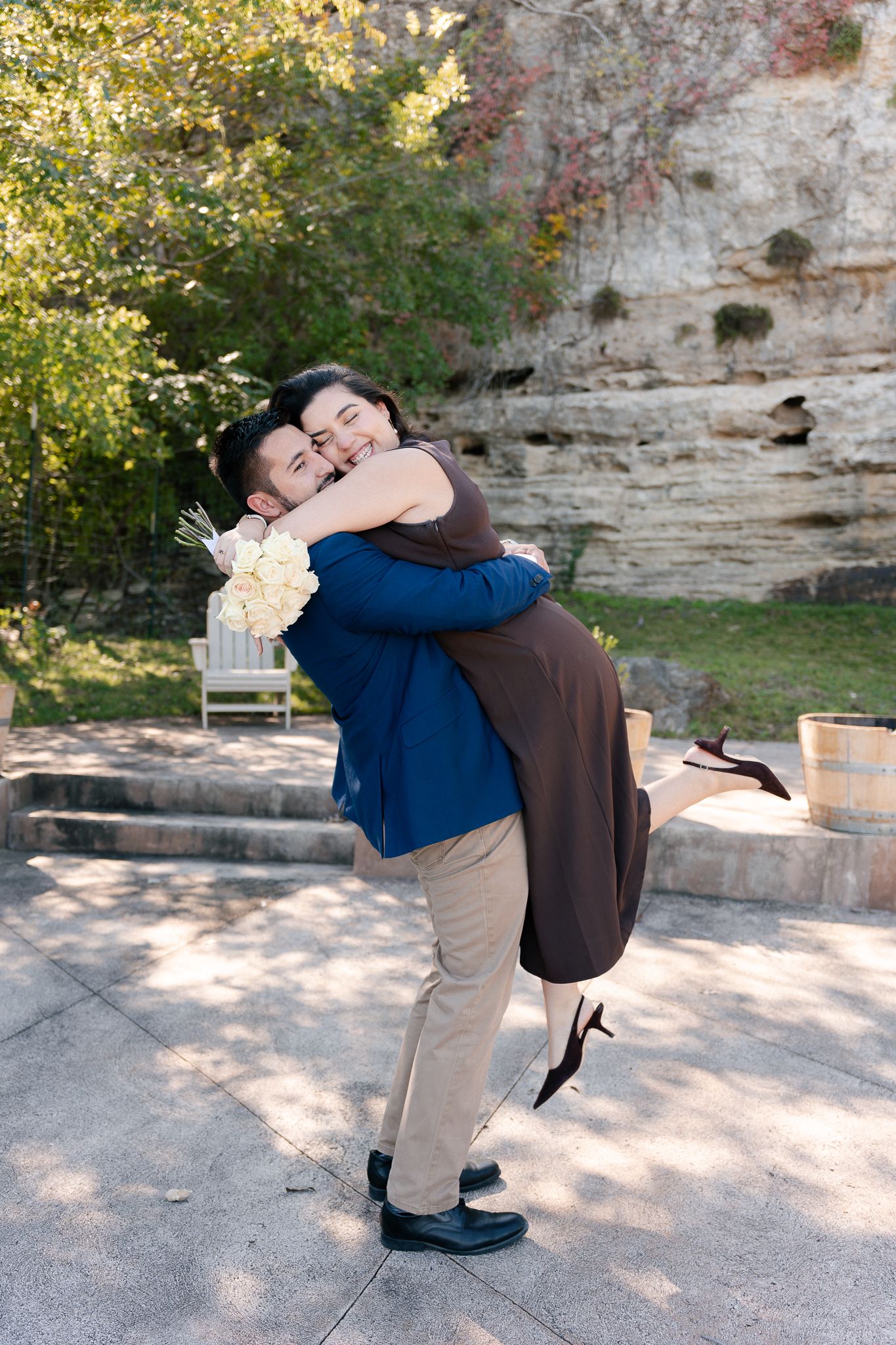 A joyful embrace captured outdoors against a limestone cliff backdrop with dappled sunlight and autumn foliage.