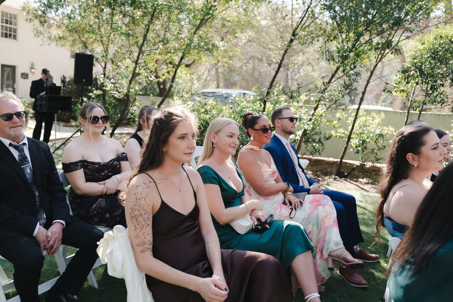 Guests seated outdoors at an elegant wedding ceremony, wearing formal attire including black and green dresses.