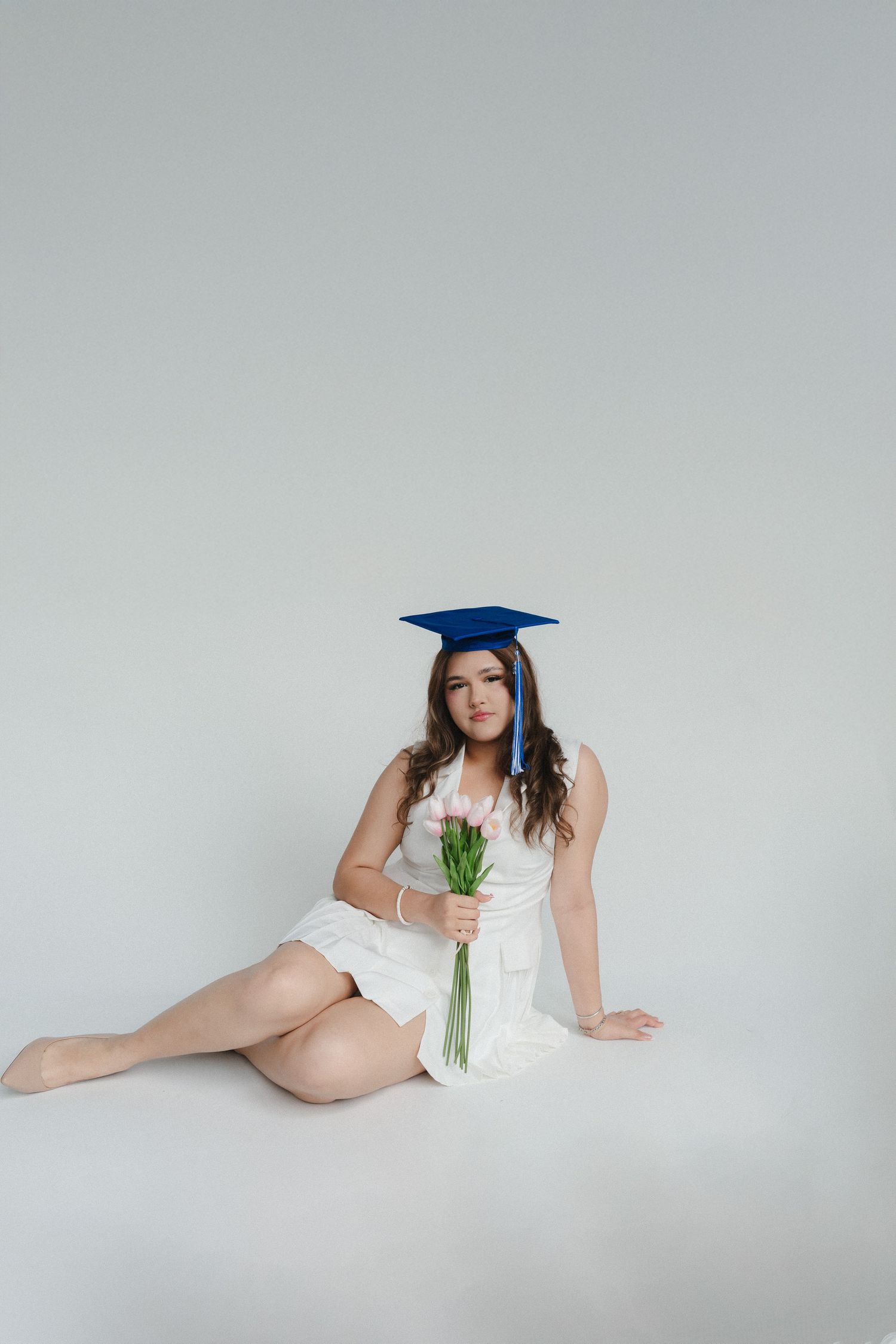 Young woman in white dress and blue graduation cap sits on floor holding pink tulips, celebrating academic achievement.