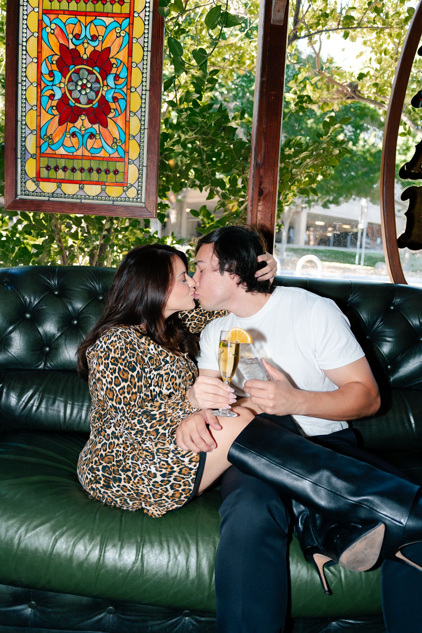 Couple sharing drinks and a kiss on a green leather couch beneath a colorful stained glass window decoration.