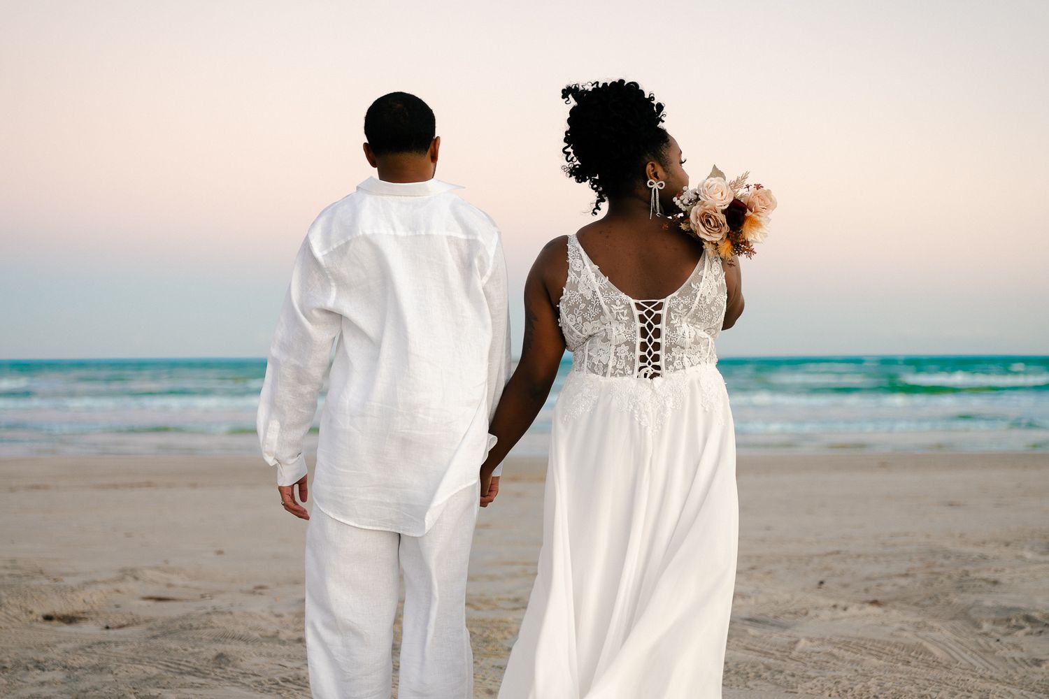 A couple in white outfits walks hand in hand along a peaceful beach at sunset.