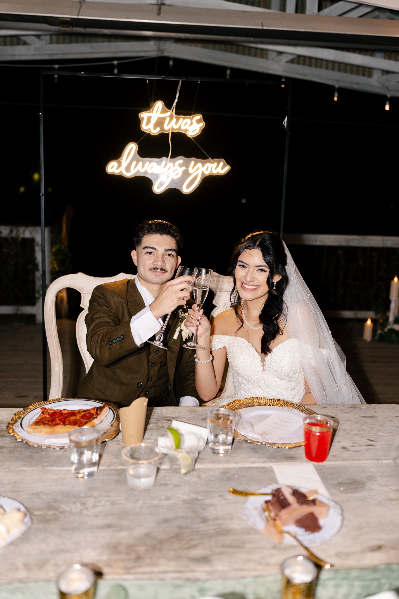 A couple enjoys their wedding reception dinner at a table with illuminated signage in the background.