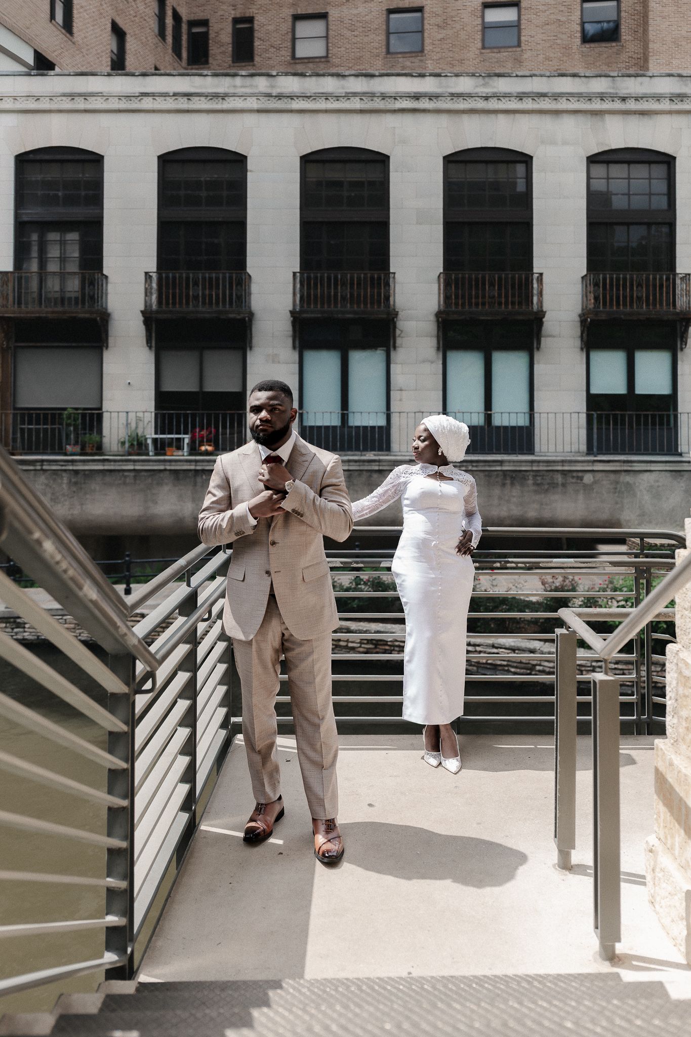 Elegant couple in beige suit and white dress posing on urban bridge with historic building backdrop.