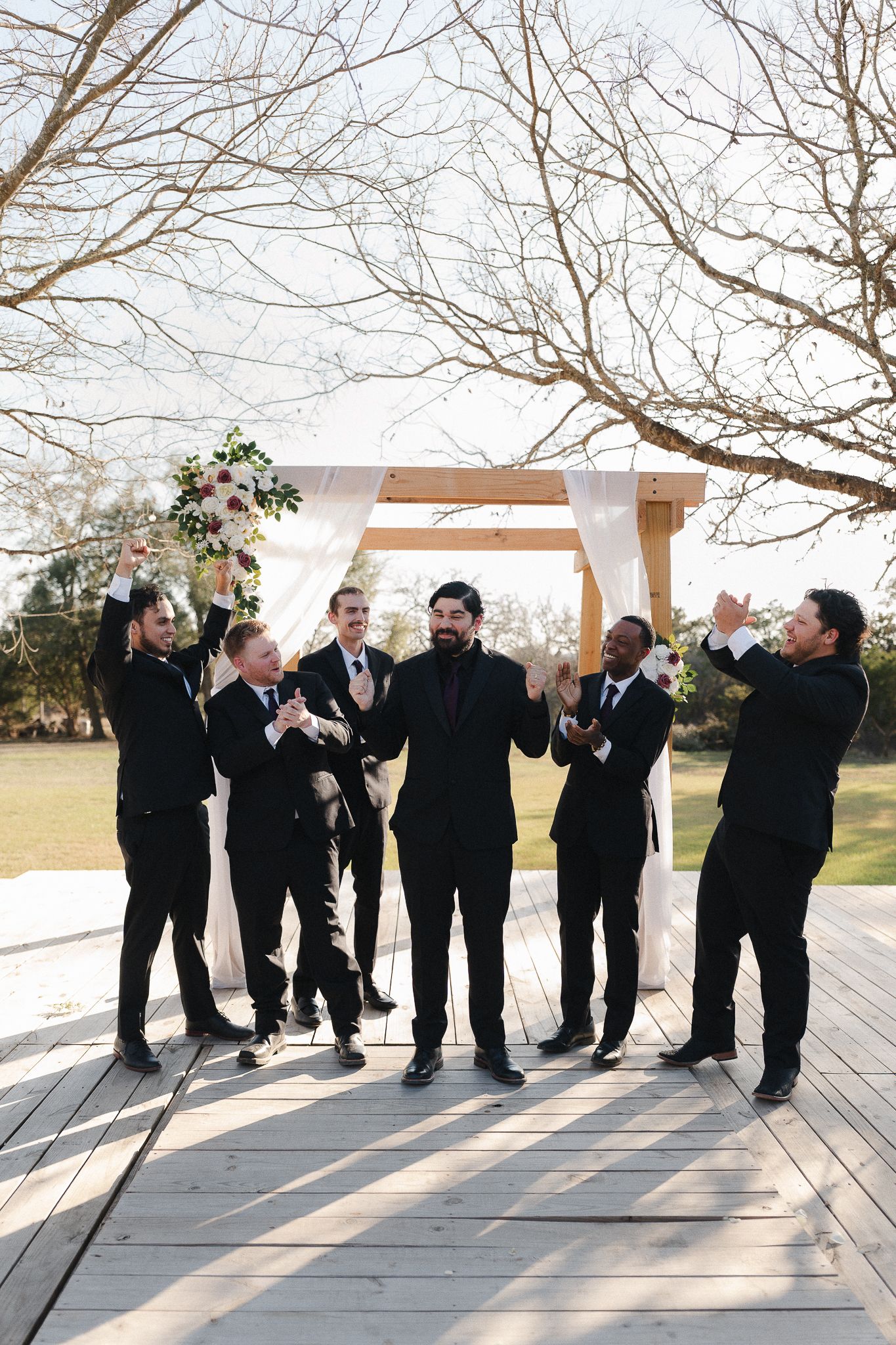 Groomsmen in black suits celebrate with the groom under a floral arch on a wooden deck at an outdoor wedding.