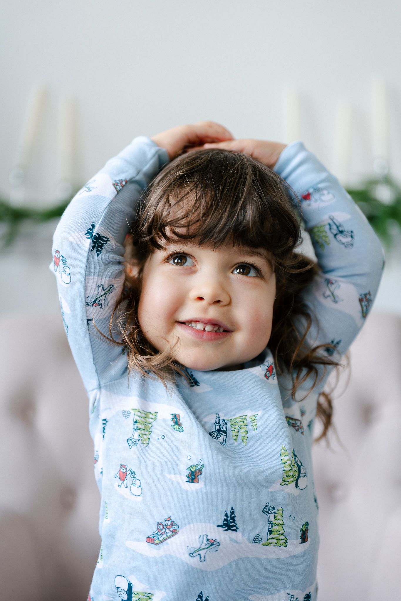 A child wearing a light blue pajama top with playful patterns stretches their arms up while smiling joyfully.