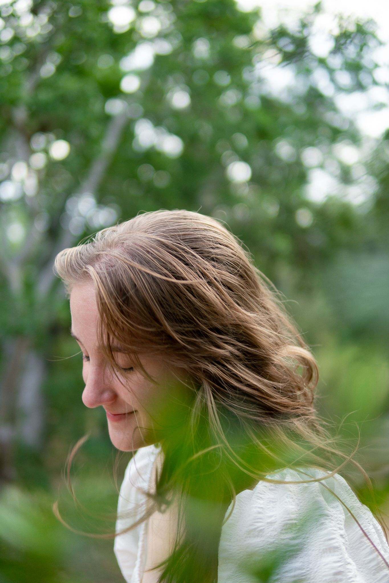 A person with flowing brown hair spins in a natural outdoor setting with green trees and bokeh lighting effects in the background.
