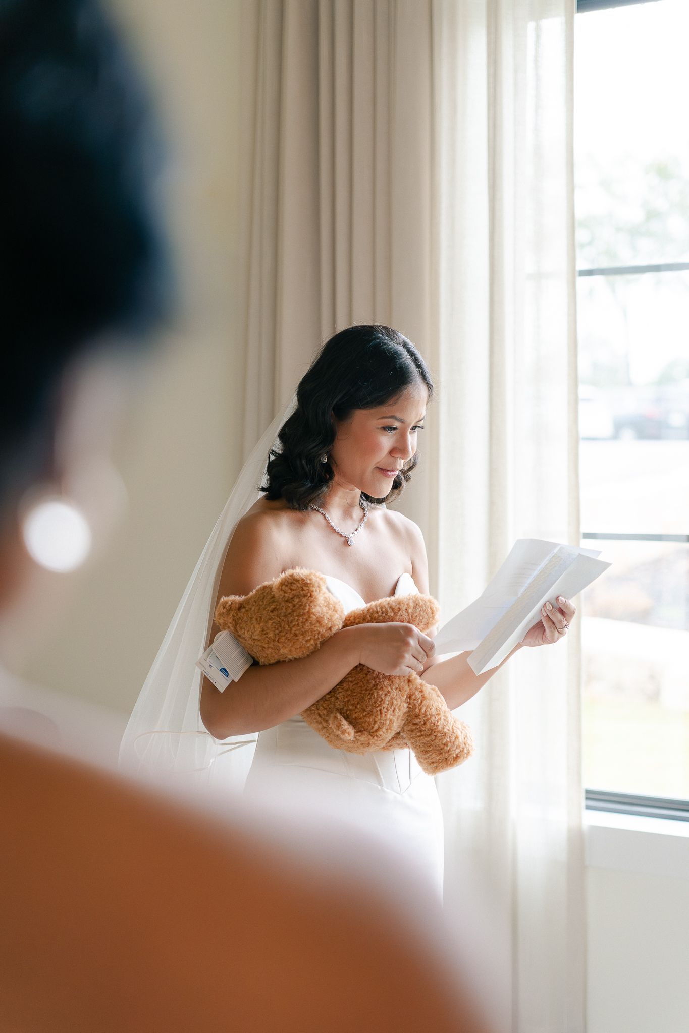 A person in a brown dress smiles joyfully while reading by a bright window in a natural light-filled room.
