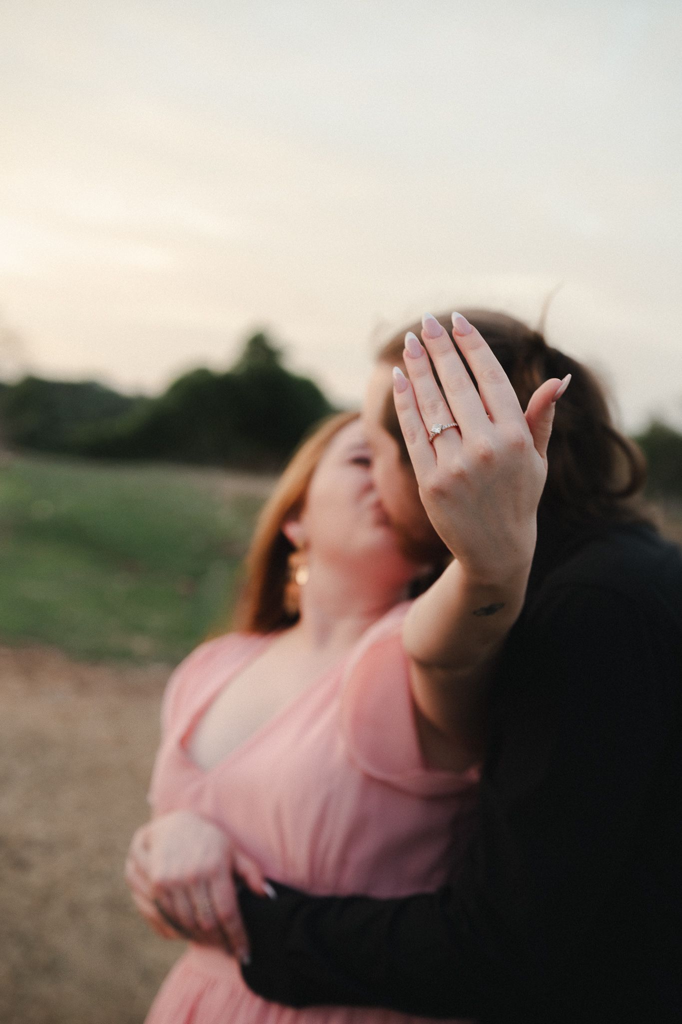 Woman showing engagement ring while kissing partner outdoors at sunset.