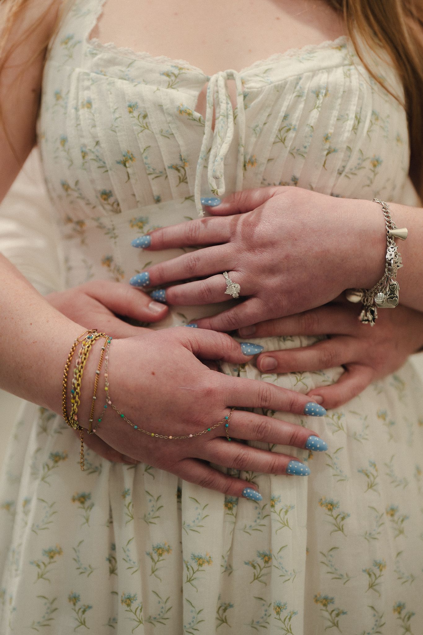 Pregnant woman in floral dress with blue acrylic nails, diamond ring, gold bracelets, and charm bracelet cradling baby bump.