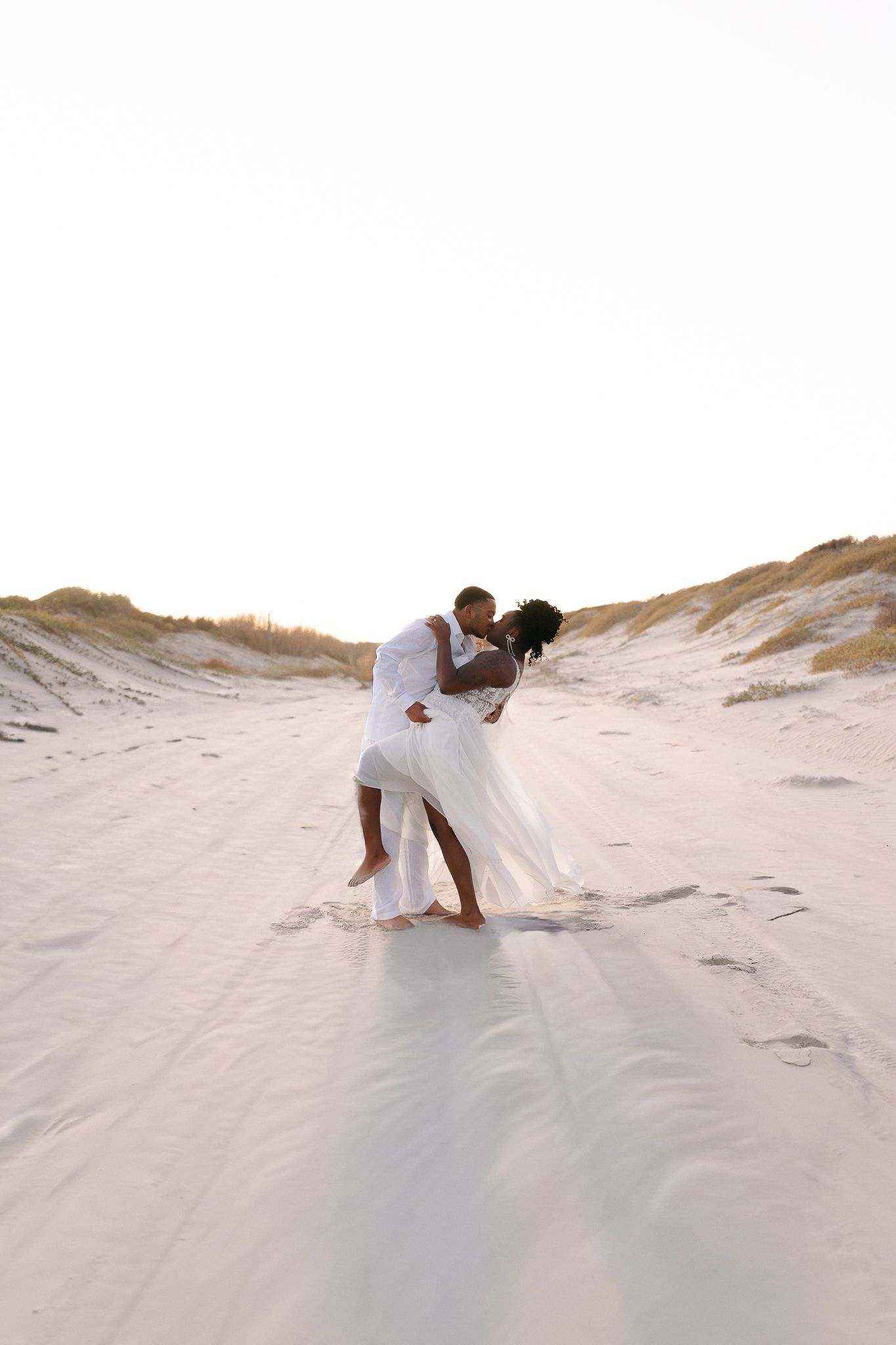 A couple in white attire sharing a romantic kiss on sandy beach dunes at sunset.