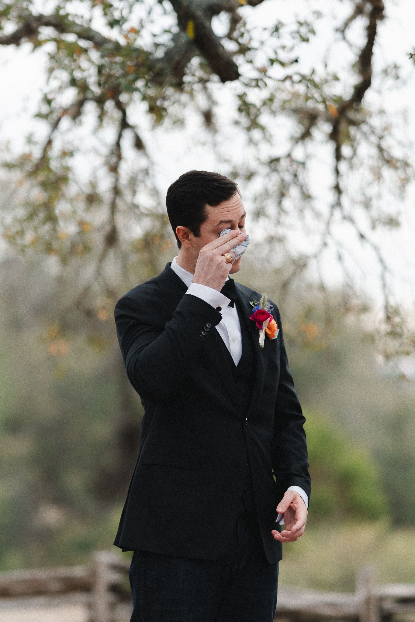 Emotional groom in black tuxedo wiping tears during wedding first look ceremony outdoors among trees.