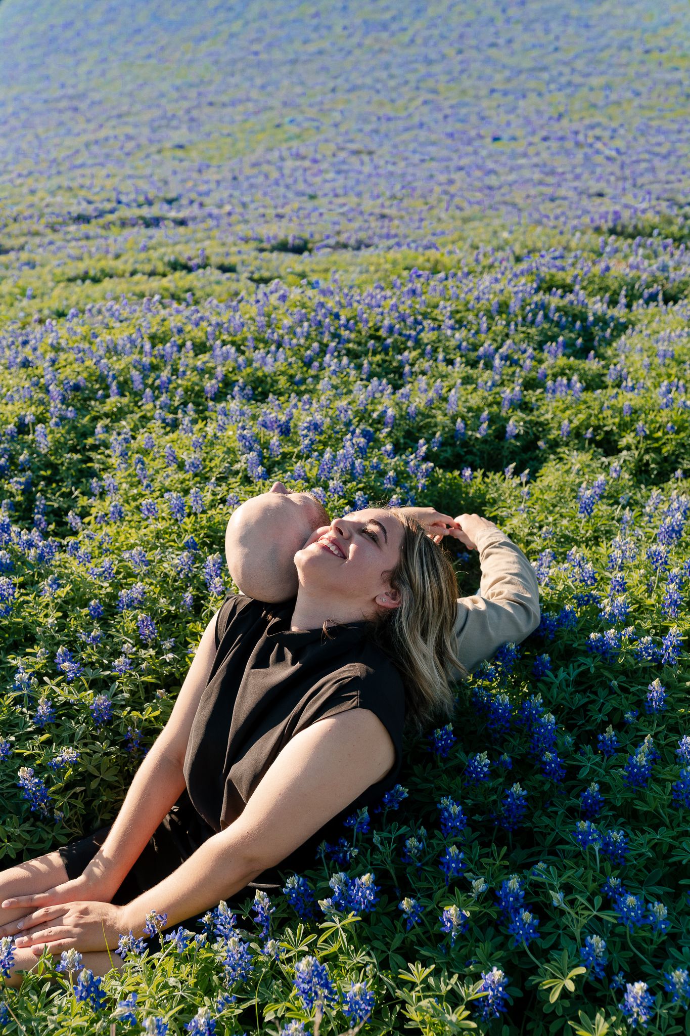 A woman lies smiling in a vibrant field of blue Texas bluebonnet wildflowers, shielding her eyes from the sun.