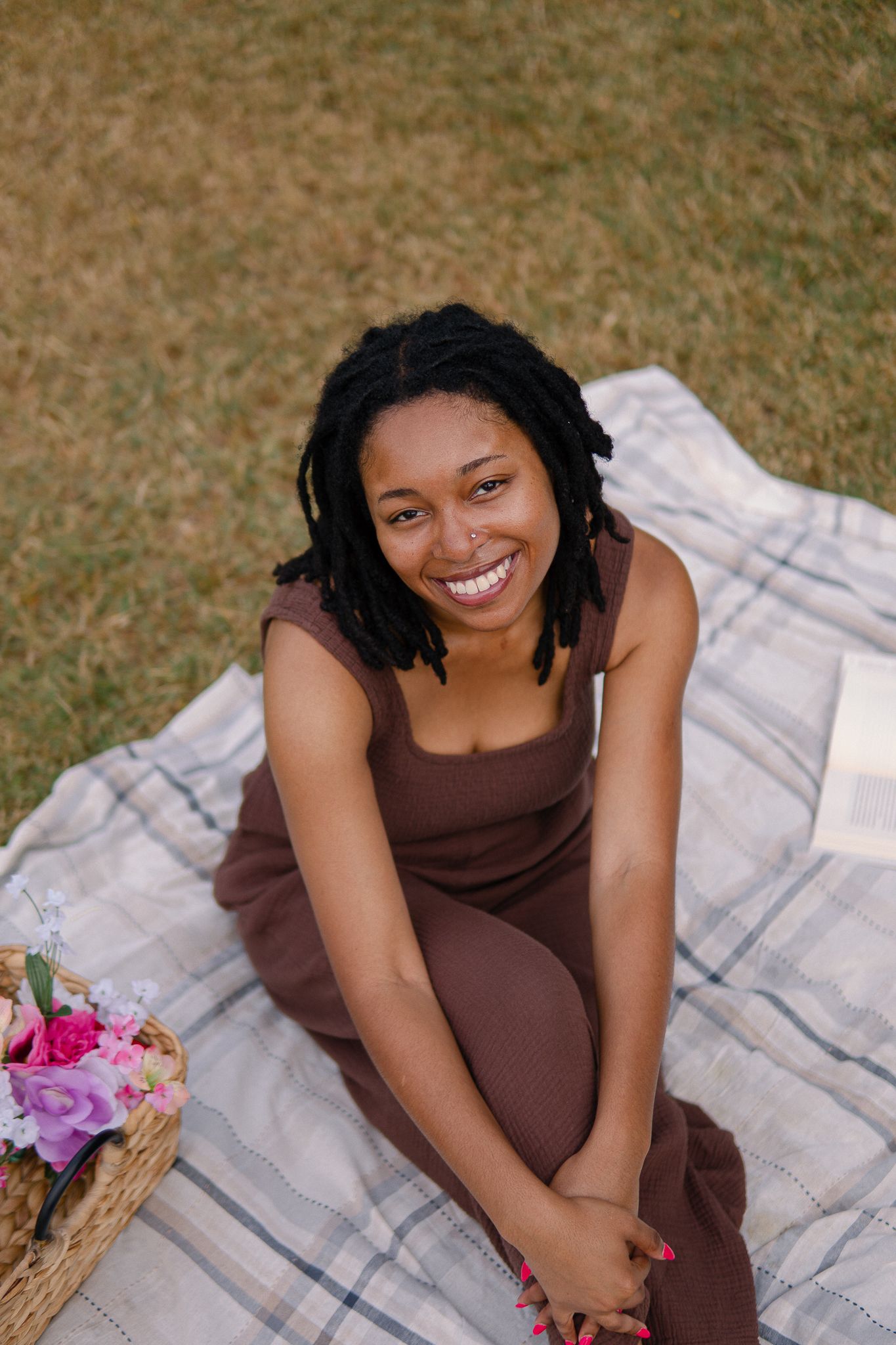 A person in a brown dress sits on a plaid blanket outdoors with colorful flowers nearby.