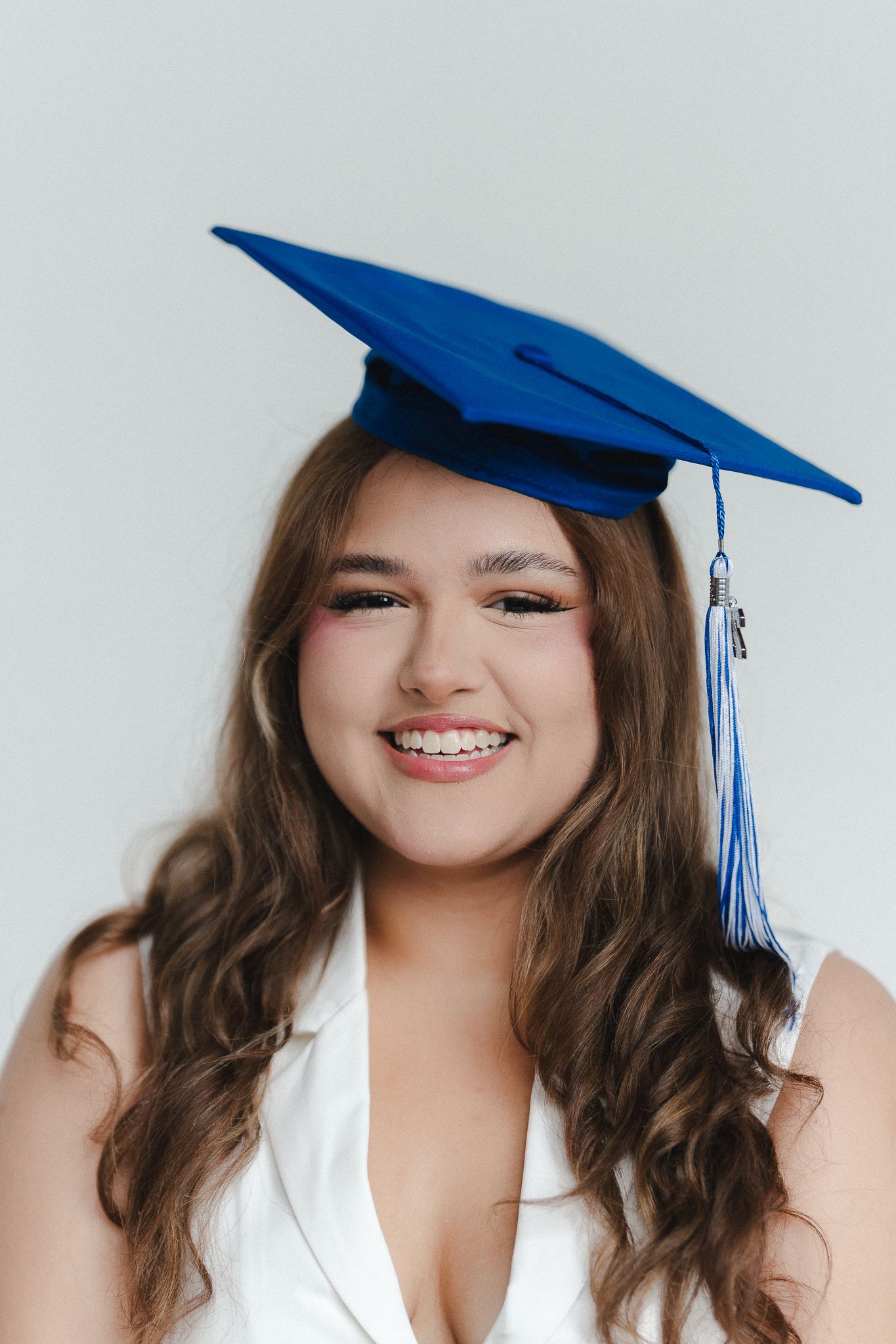 Smiling young woman wearing a blue graduation cap with tassel, long wavy hair, white outfit, light background.