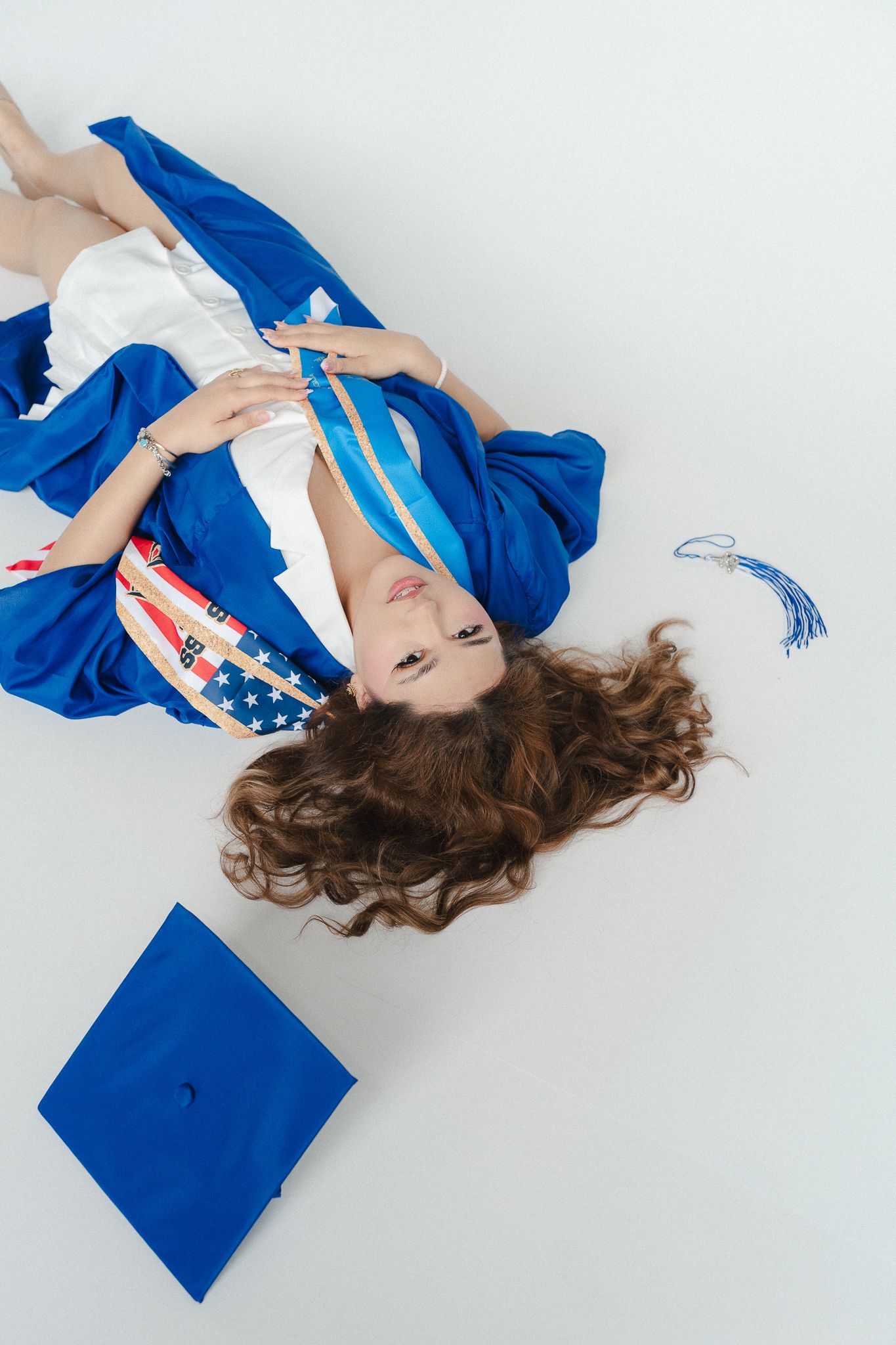 Young woman in blue graduation gown lying on white floor with mortarboard cap and American flag sash.