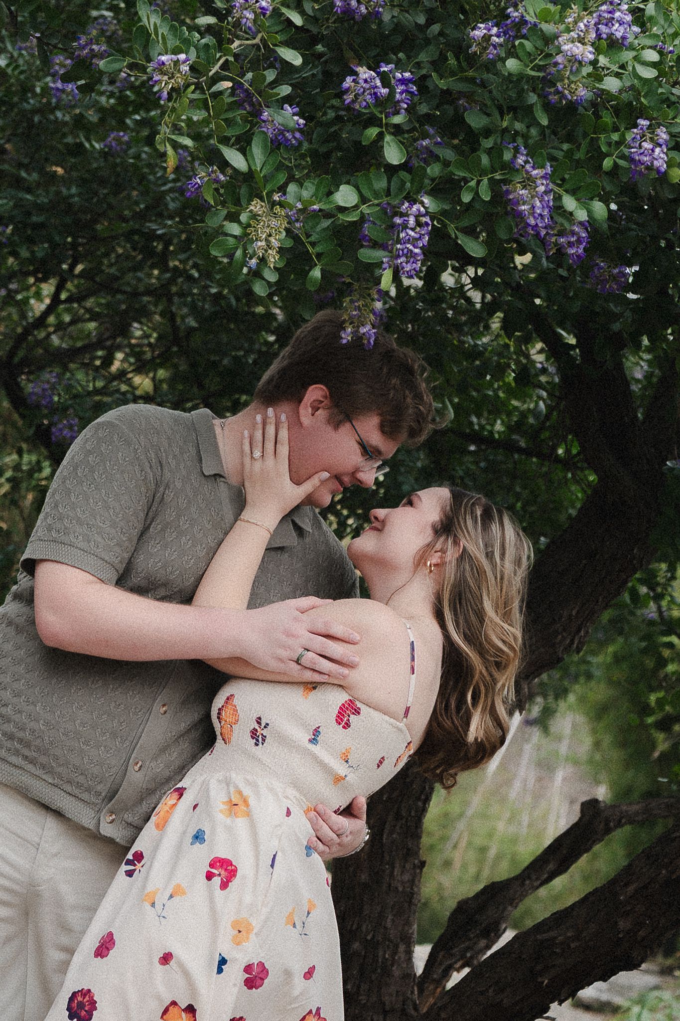 Couple sharing a romantic moment under a blooming purple flower tree, woman in floral dress, man in olive shirt.