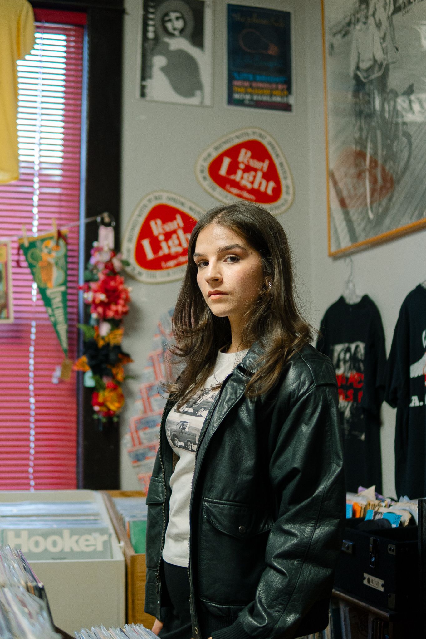 A person in a black leather jacket sits in a room decorated with posters and red window blinds.